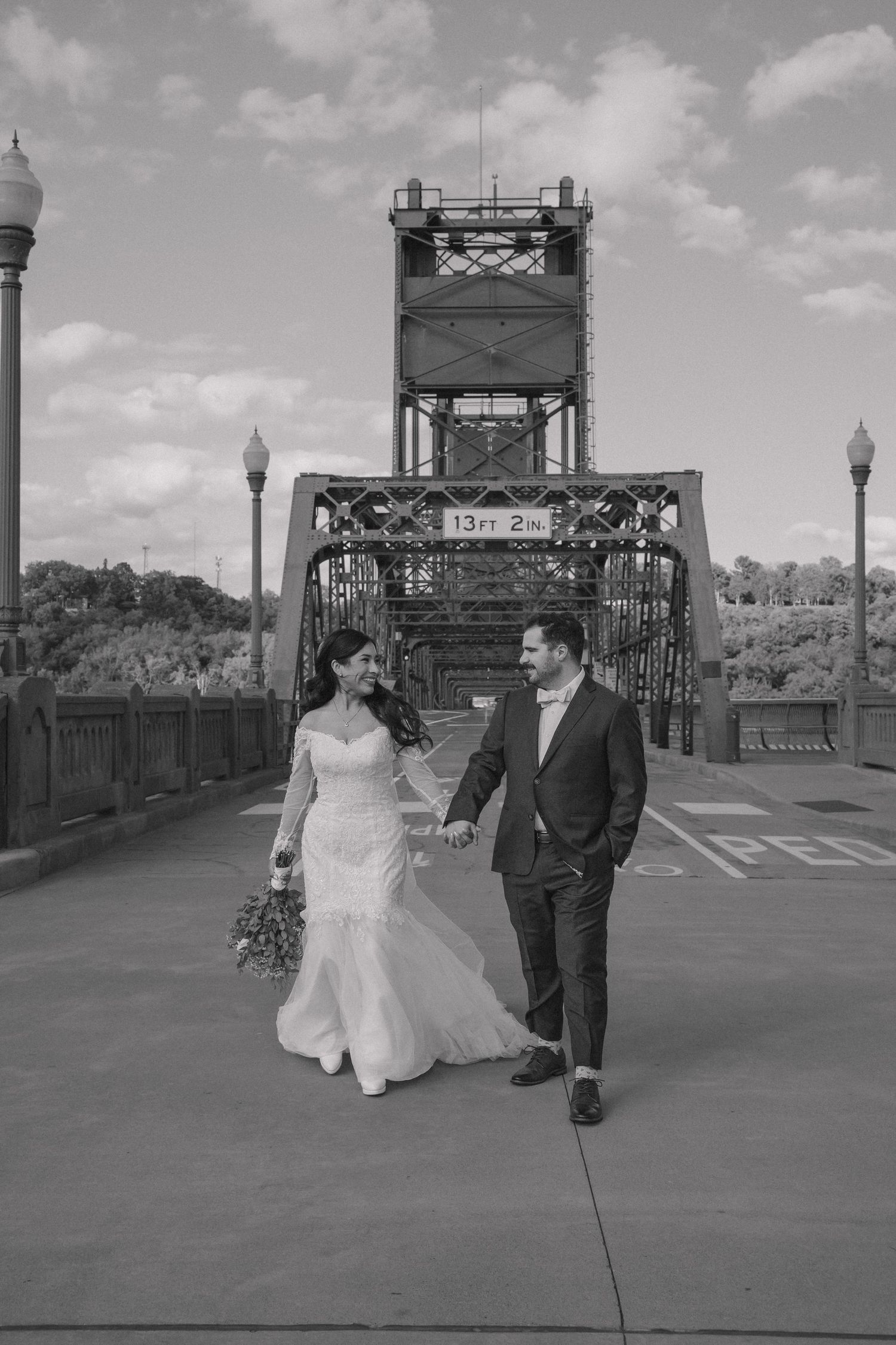 Black and white photo of bride and groom walking hand in hand on bridge.