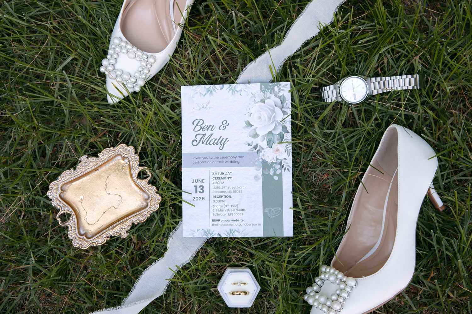 Wedding flat lay with floral invitation, white heels, pearl shoes, gold ring box, watch, and ribbon on green grass.