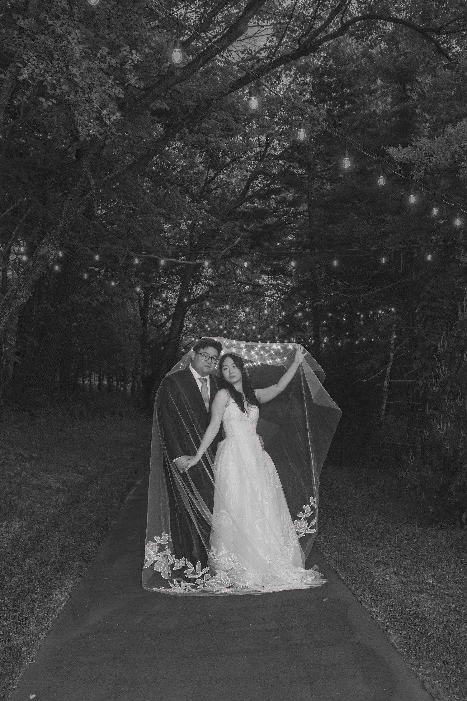 Black and white wedding photo of couple under string lights in forest, bride in gown with floral veil, groom in suit.