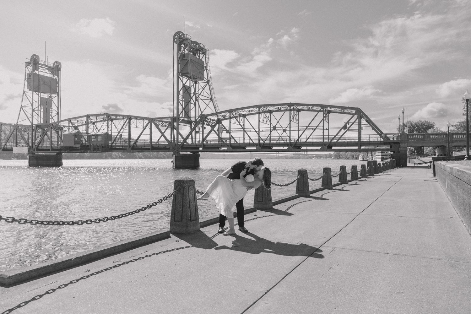 Black and white photo of a couple sharing a romantic moment on a waterfront promenade near a historic lift bridge.