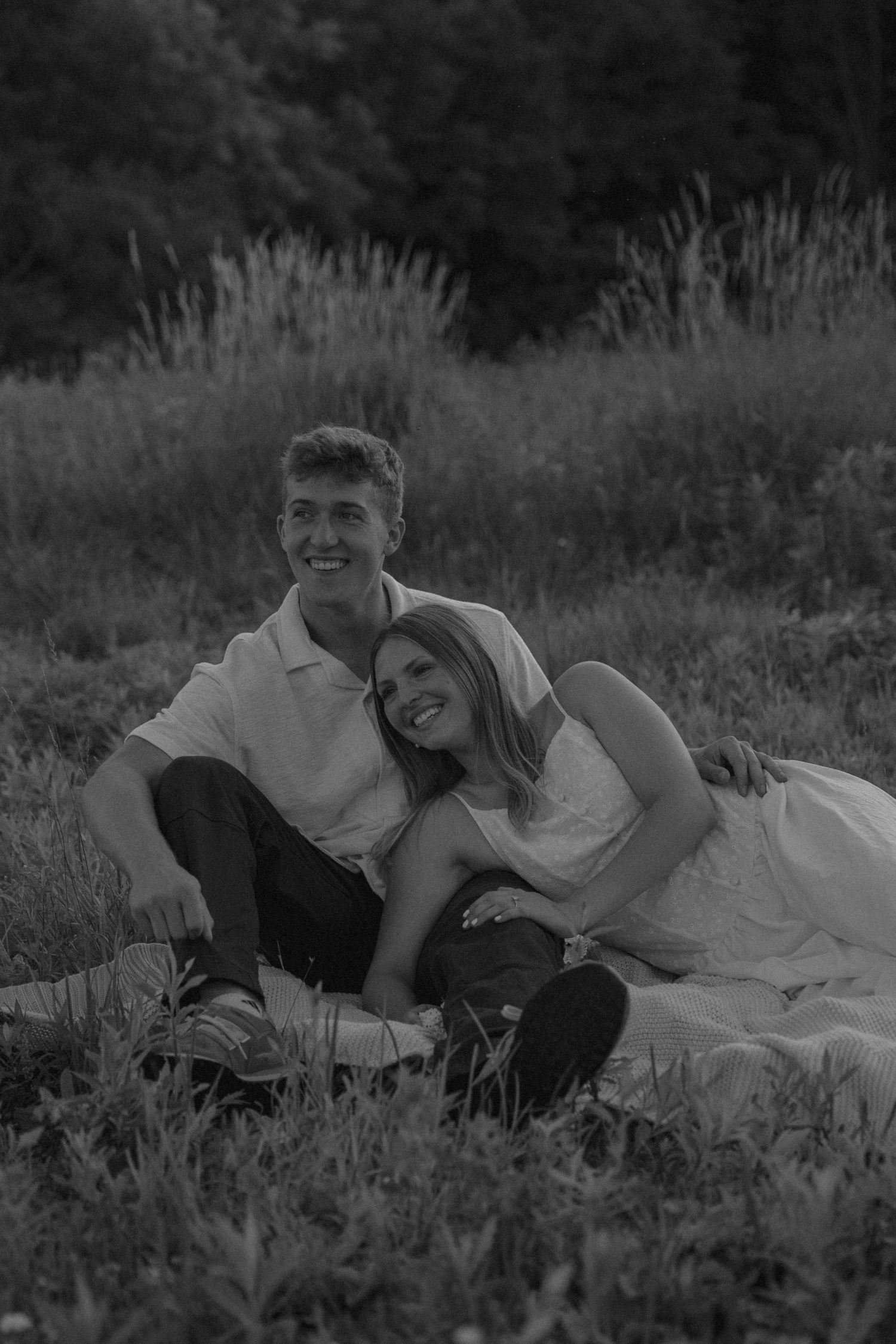 Black and white photo of a smiling couple sitting together in a grassy field, leaning close in a romantic outdoor setting.