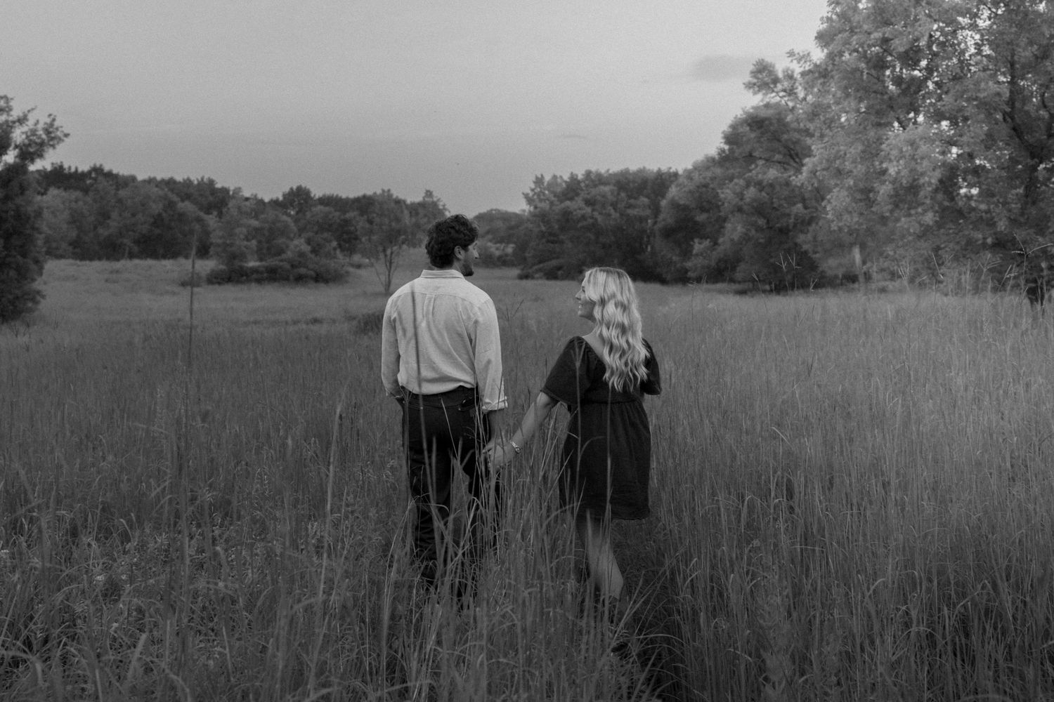 Black and white photo of couple holding hands walking through tall grass field at dusk.