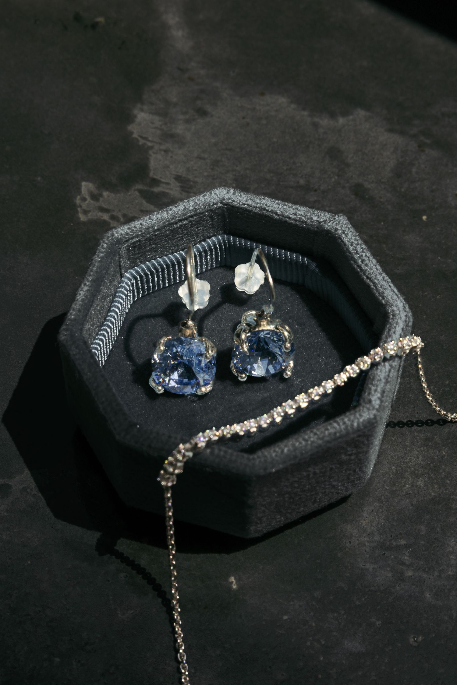 Blue gemstone drop earrings and diamond tennis bracelet displayed in a dark hexagonal velvet jewelry box.