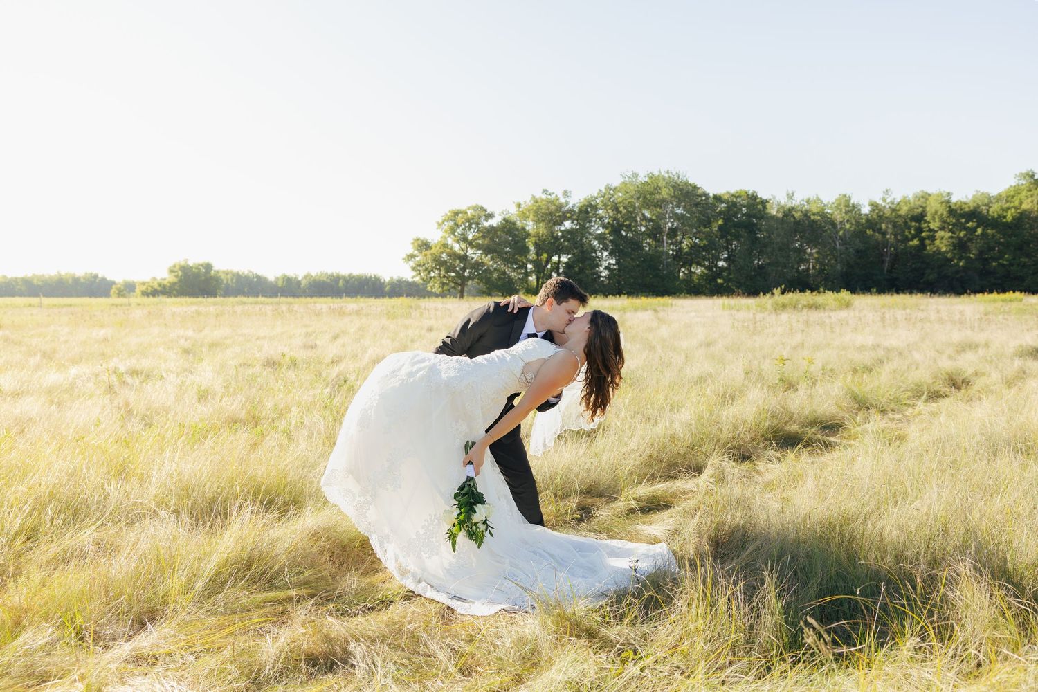 Groom dipping bride for a kiss in golden sunlit open field on wedding day.