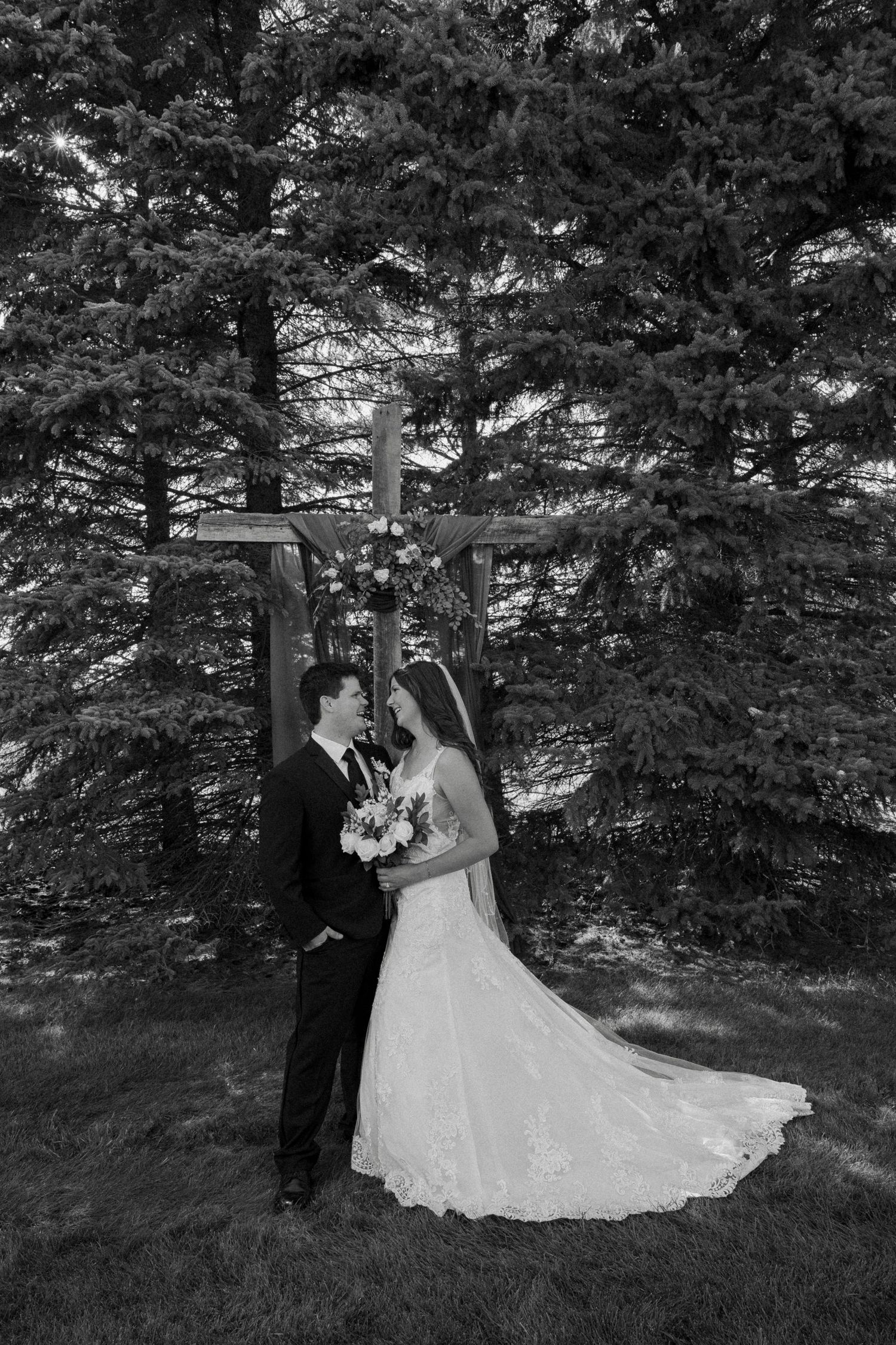 Bride and groom share a romantic moment under a wooden cross surrounded by evergreen trees in a black and white wedding photo.