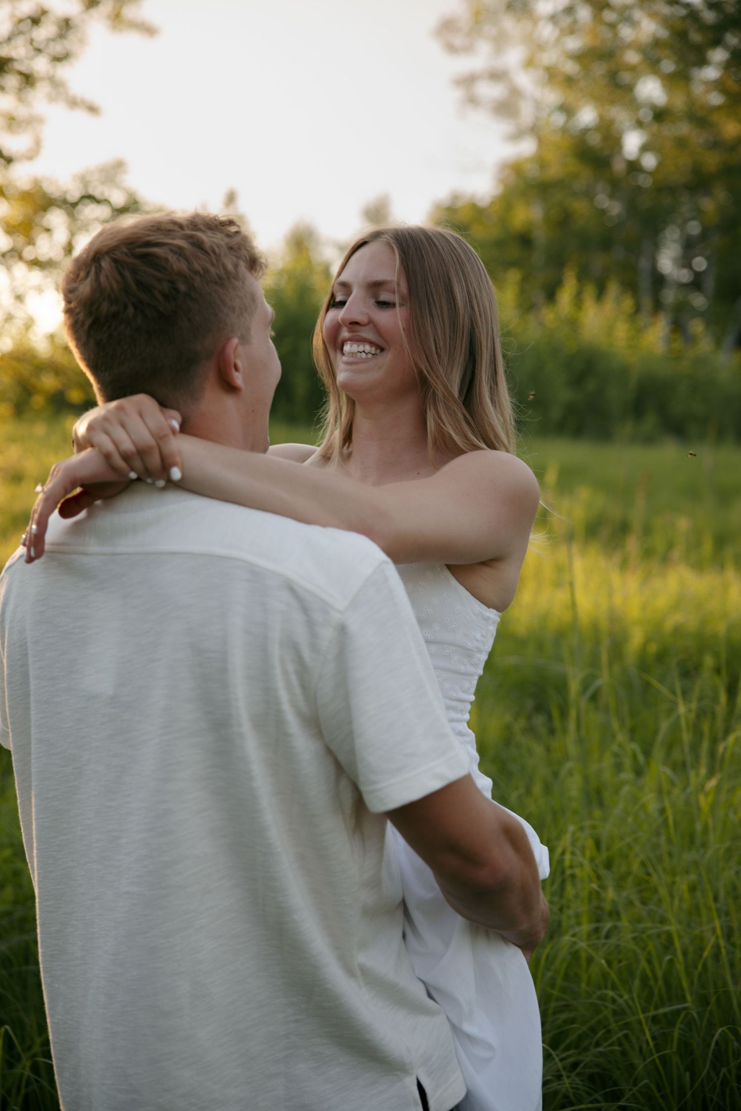 A smiling woman in a white dress embraces a man in a green field during golden hour sunset.