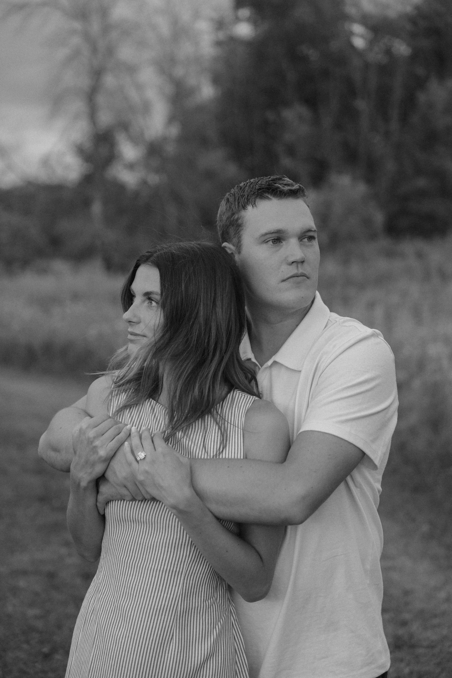 Black and white photo of a young couple embracing outdoors, man hugging woman from behind in a field.