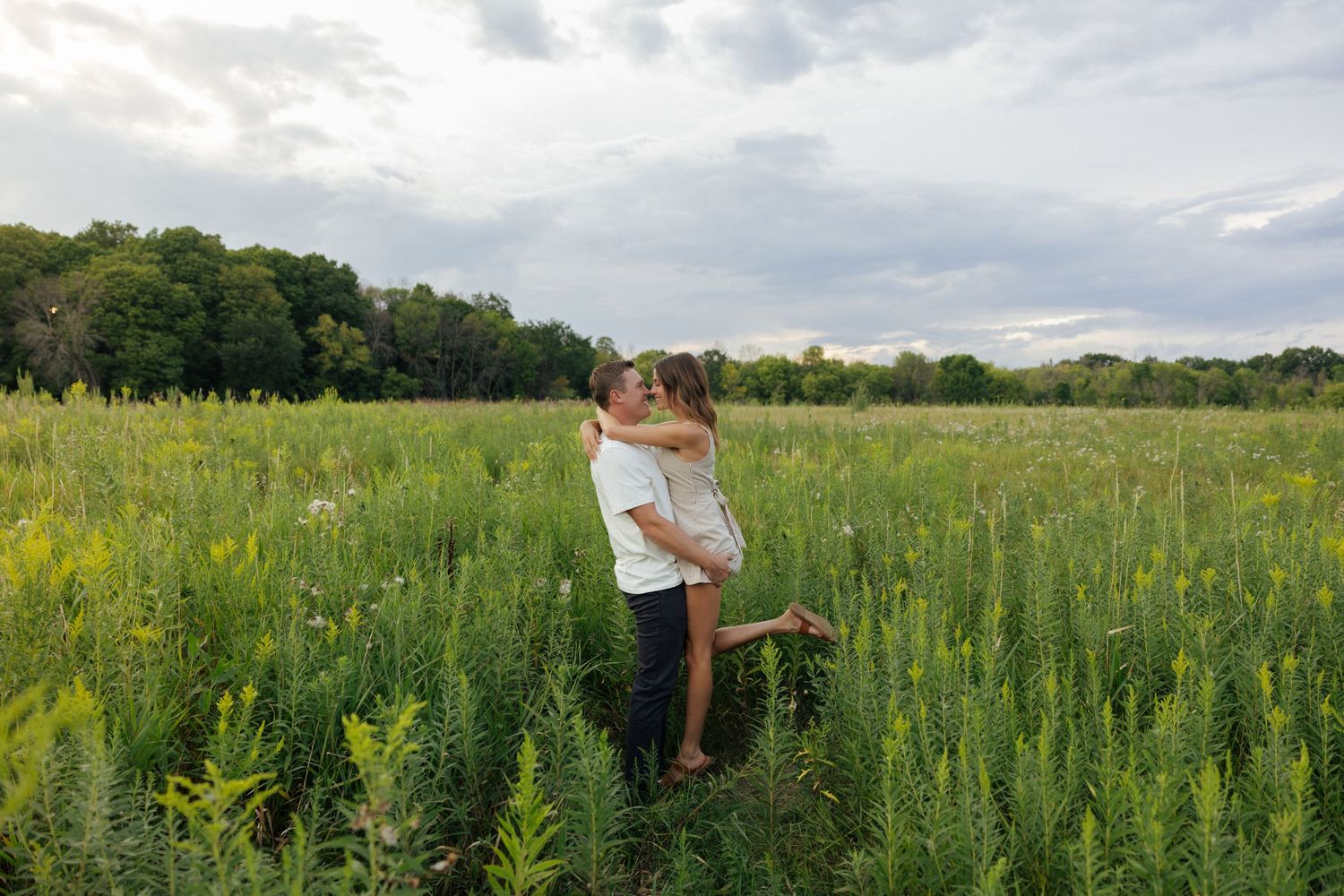 Man lifting woman in an embrace in a vibrant green meadow with wildflowers under a cloudy sky.