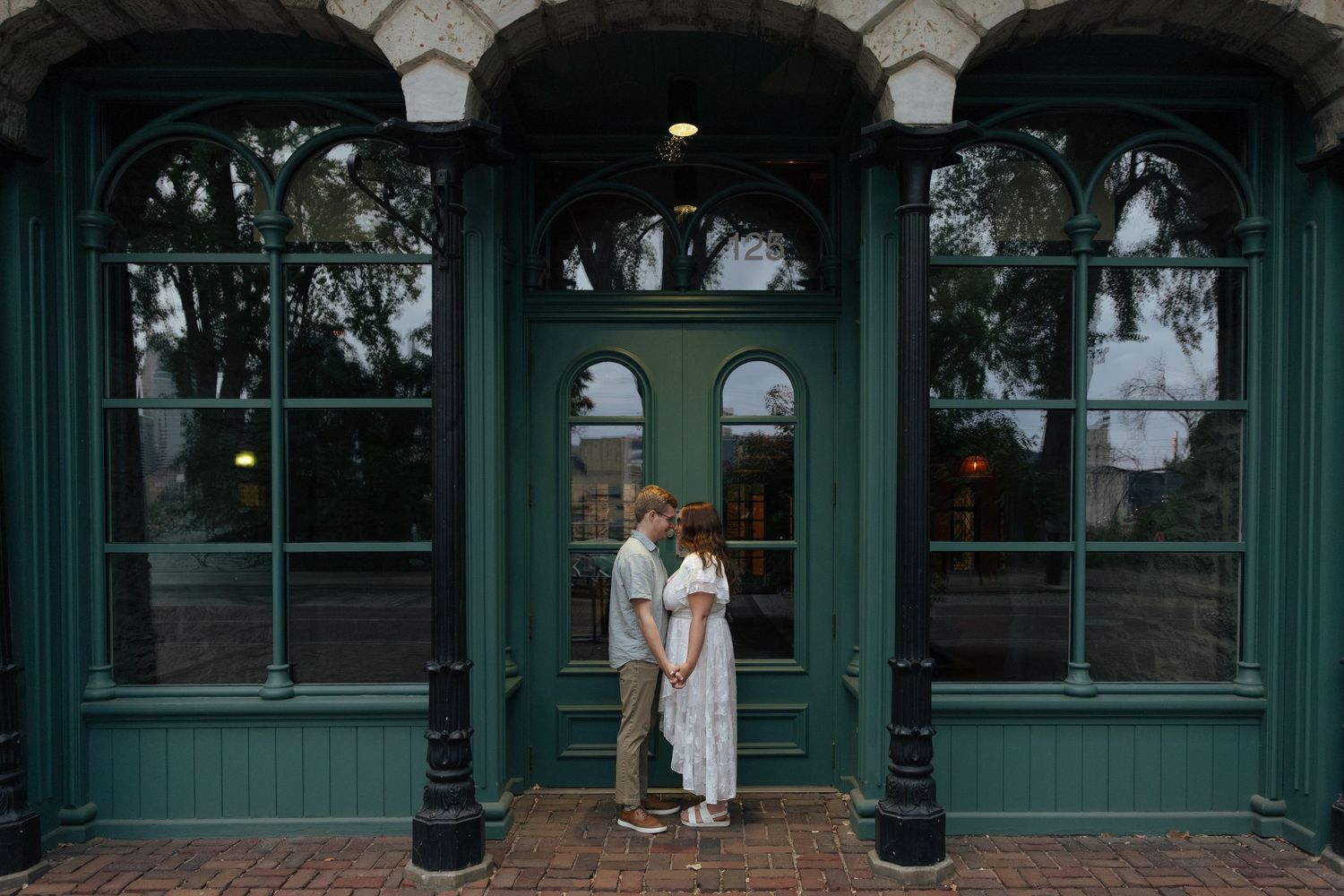 Couple embracing in front of elegant green Victorian building with arched windows and doors.
