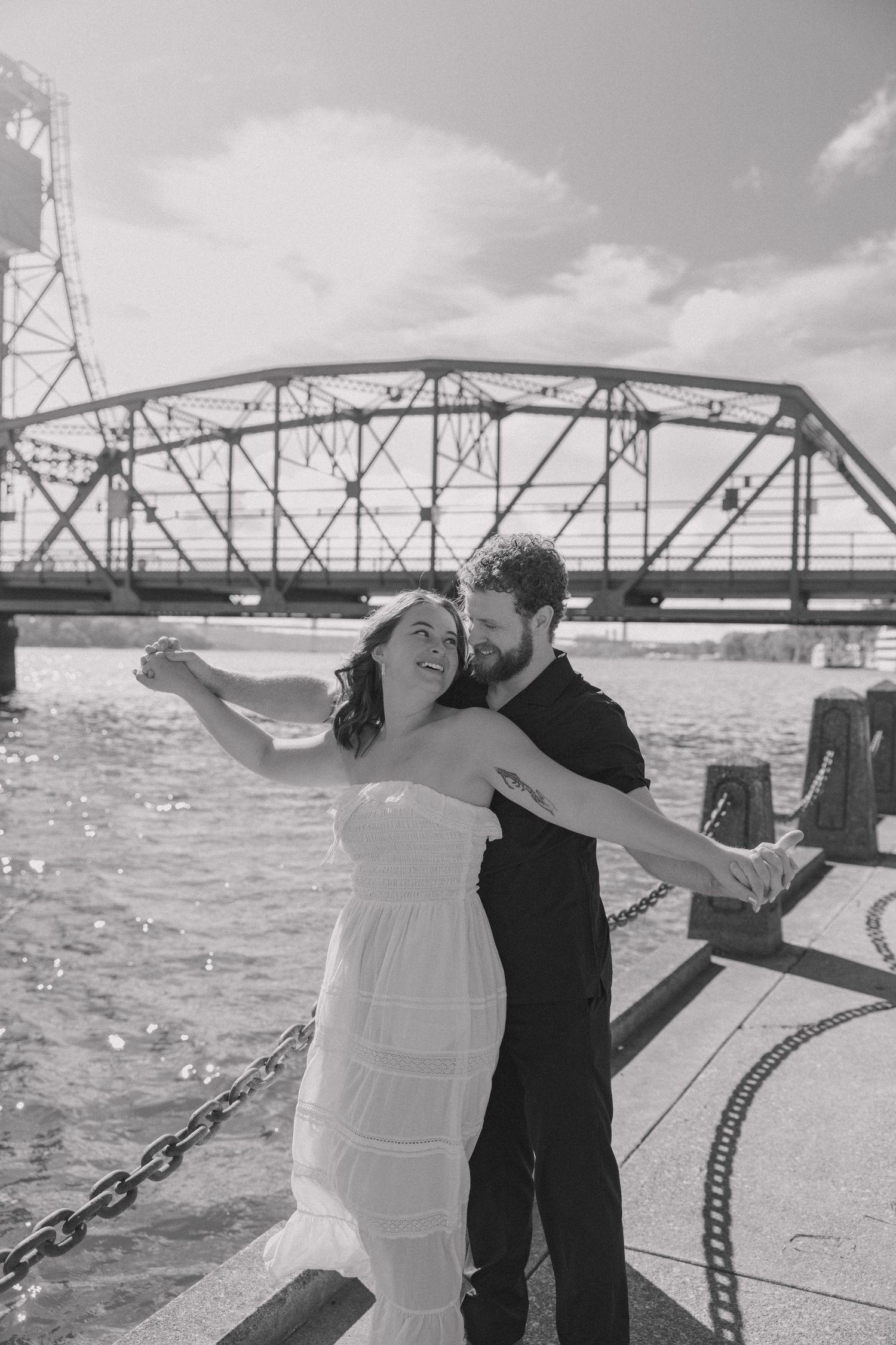 Bride and groom dancing romantically by the river with a steel swing bridge in black and white.