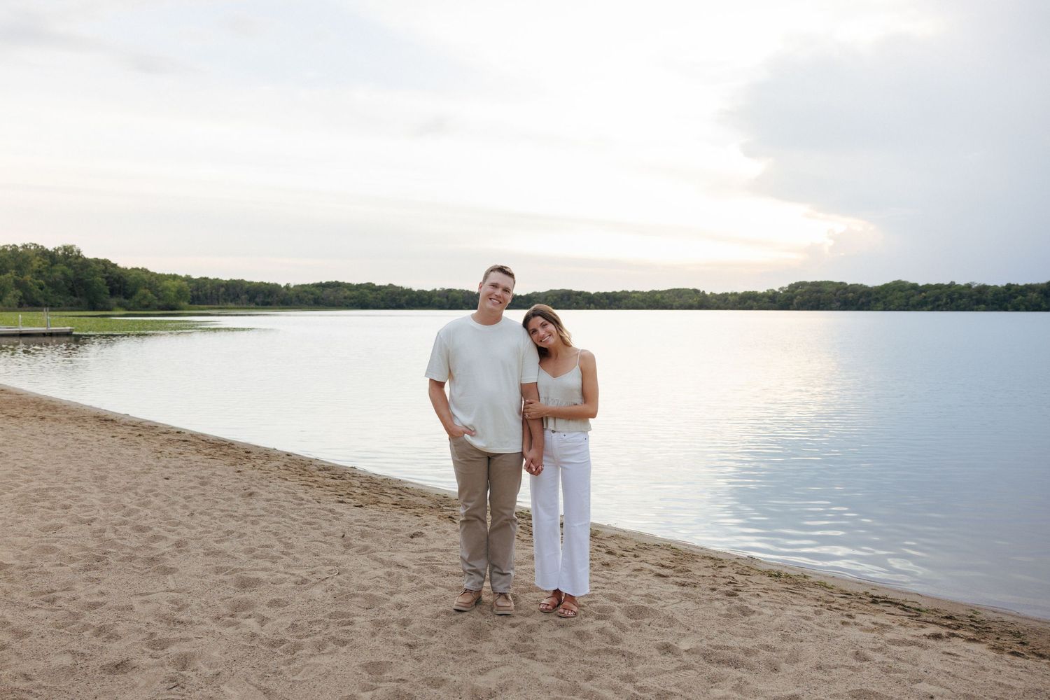 Smiling couple standing together on a sandy beach at sunset with calm lake behind them.