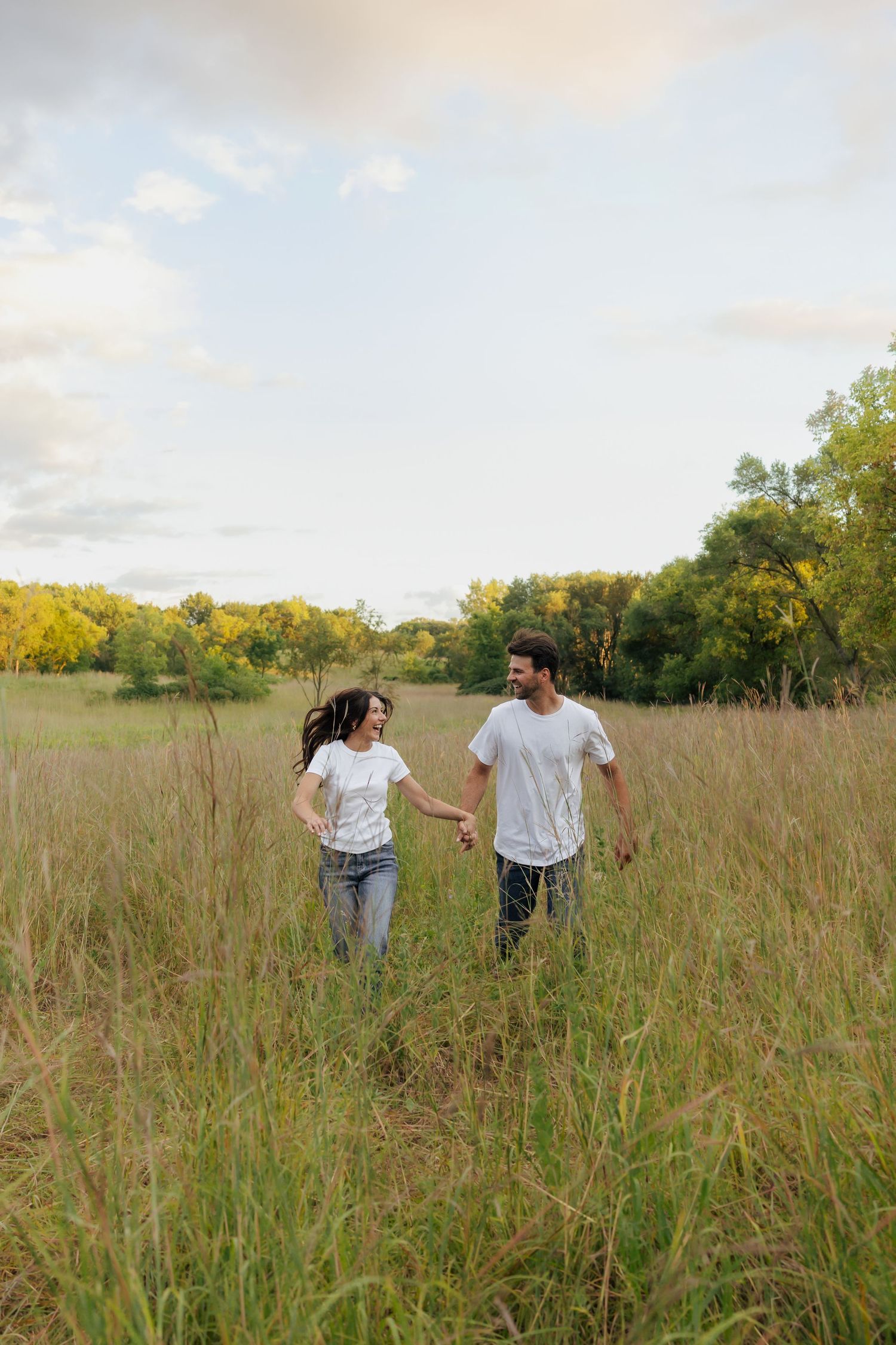 Happy couple holding hands and walking through tall grass meadow, both wearing white t-shirts and jeans.