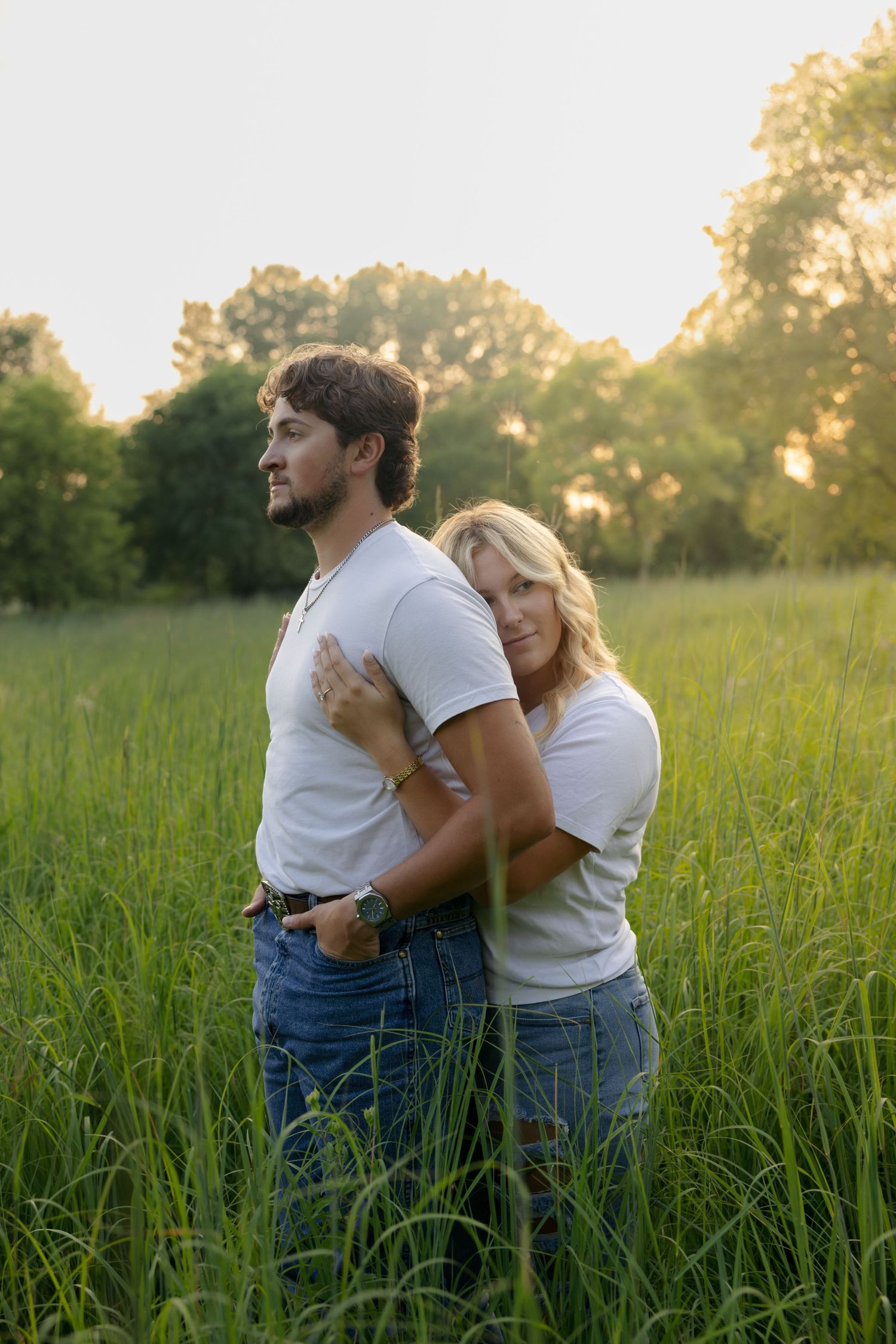 A couple in matching white tees and jeans embraces in a lush green field at golden hour.