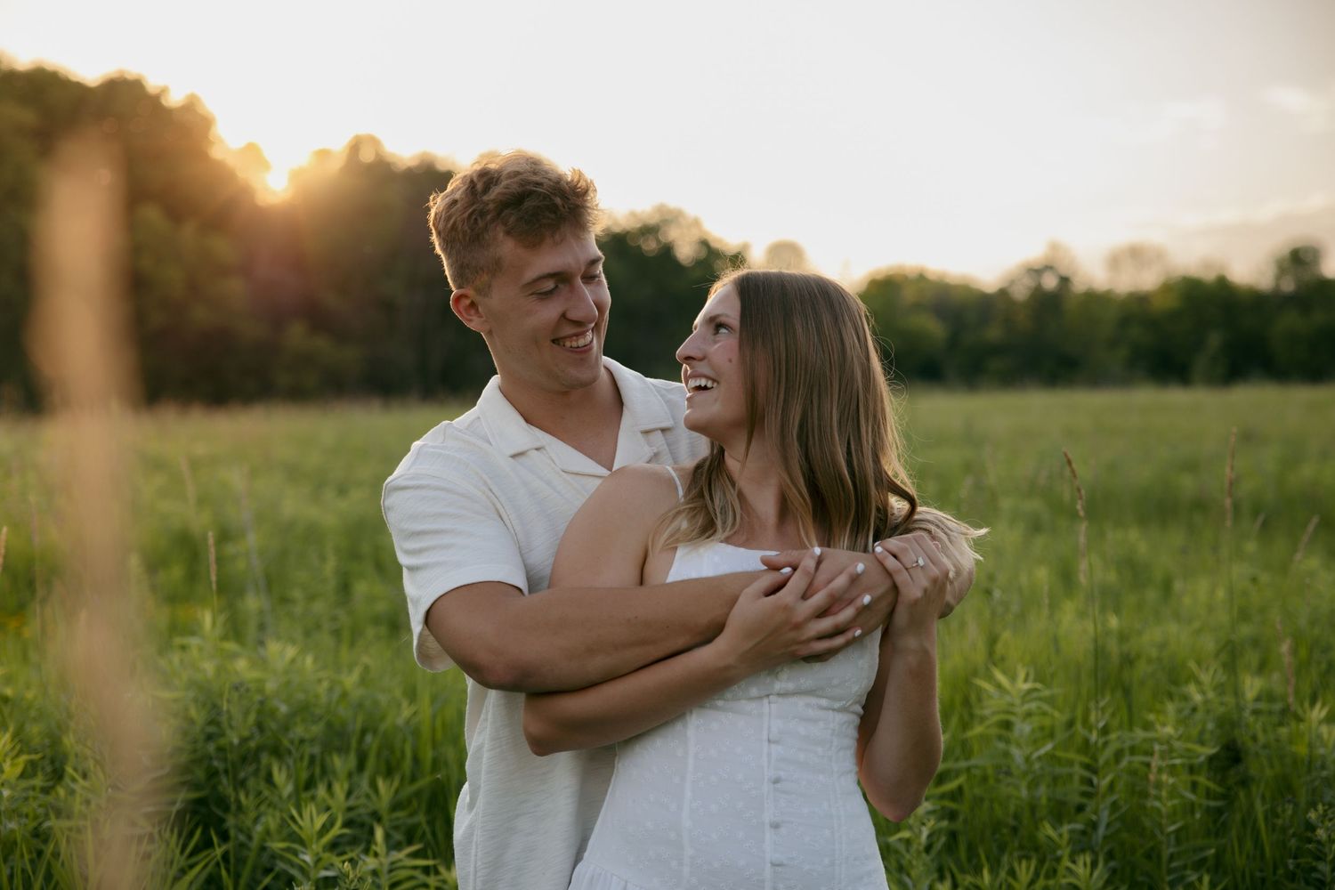 Young couple smiling and embracing in a sunlit green field during golden hour sunset outdoors.