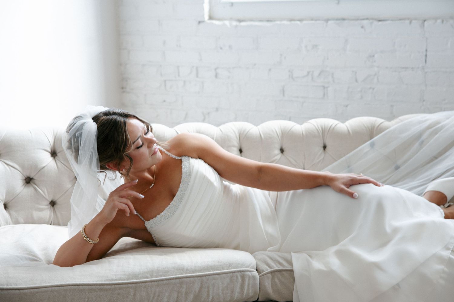 Bride in white wedding dress and veil reclining gracefully on a tufted ivory sofa in a bright room.