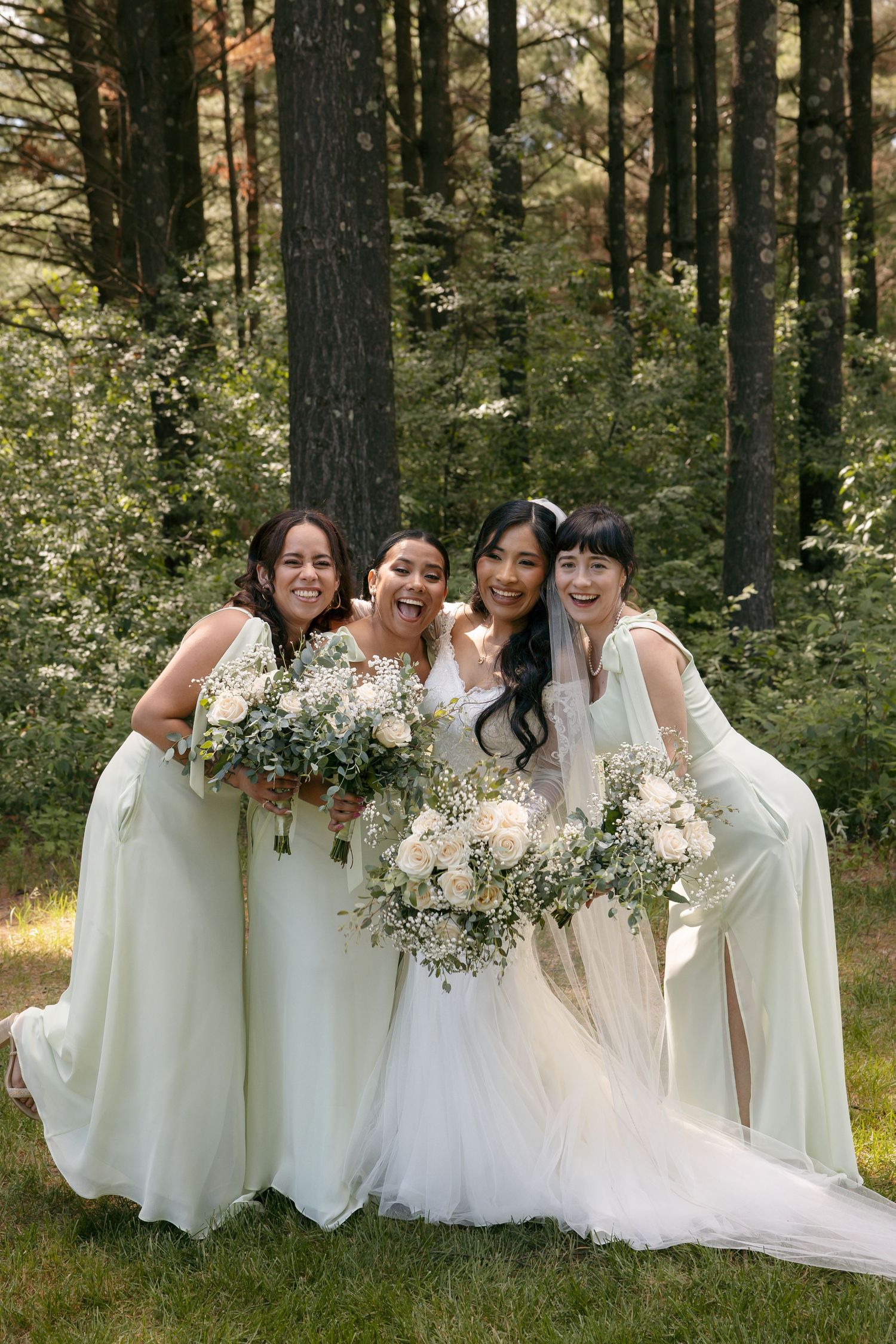 Bride in white gown poses with three bridesmaids in sage green dresses holding white floral bouquets in a forest setting.