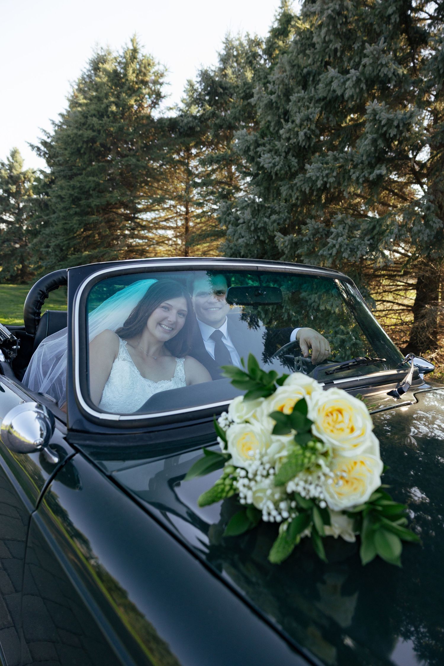 Newlyweds smiling in vintage convertible car with white rose bouquet on hood.