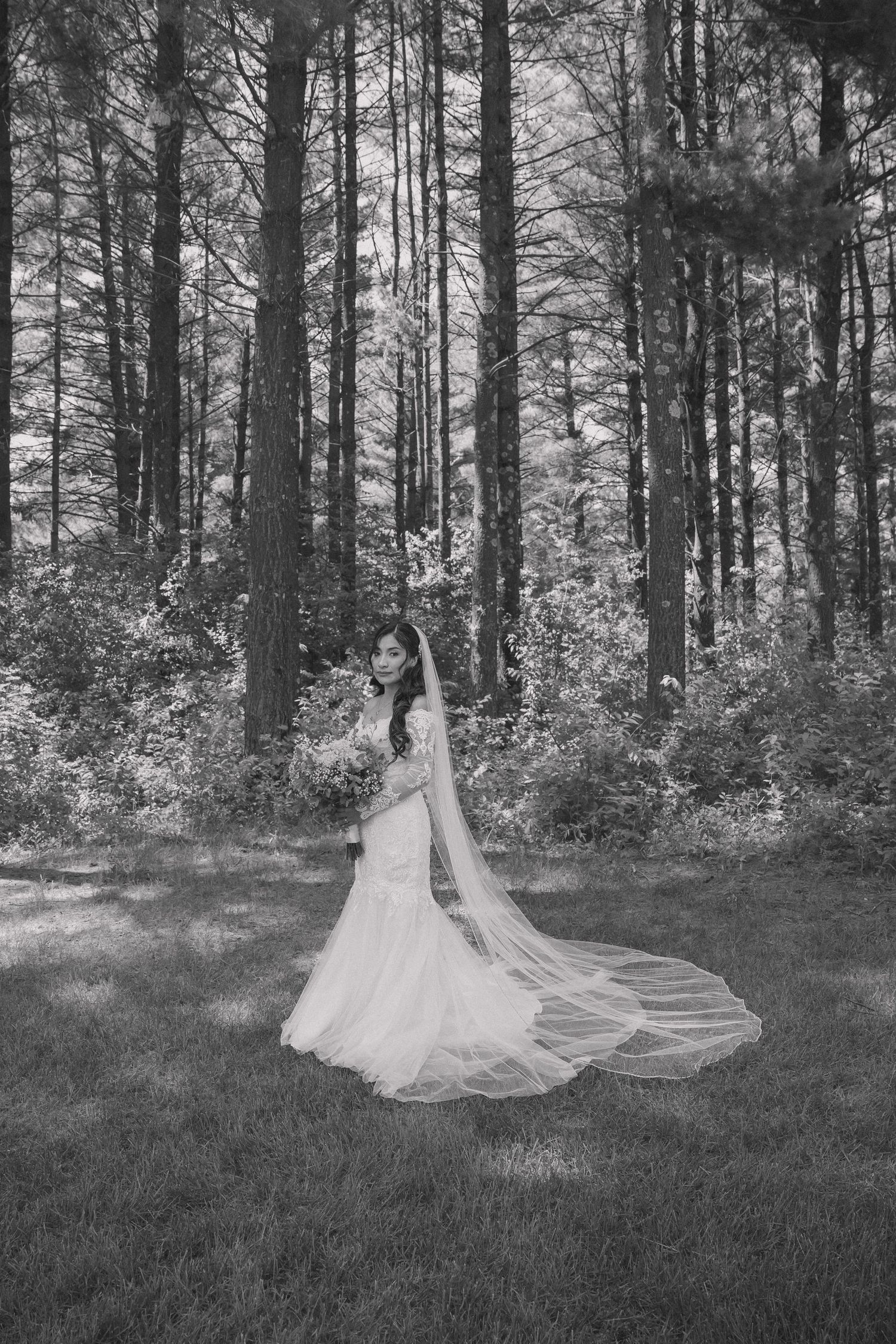 Black and white photo of a bride in a flowing gown and long veil standing gracefully in a lush forest setting.