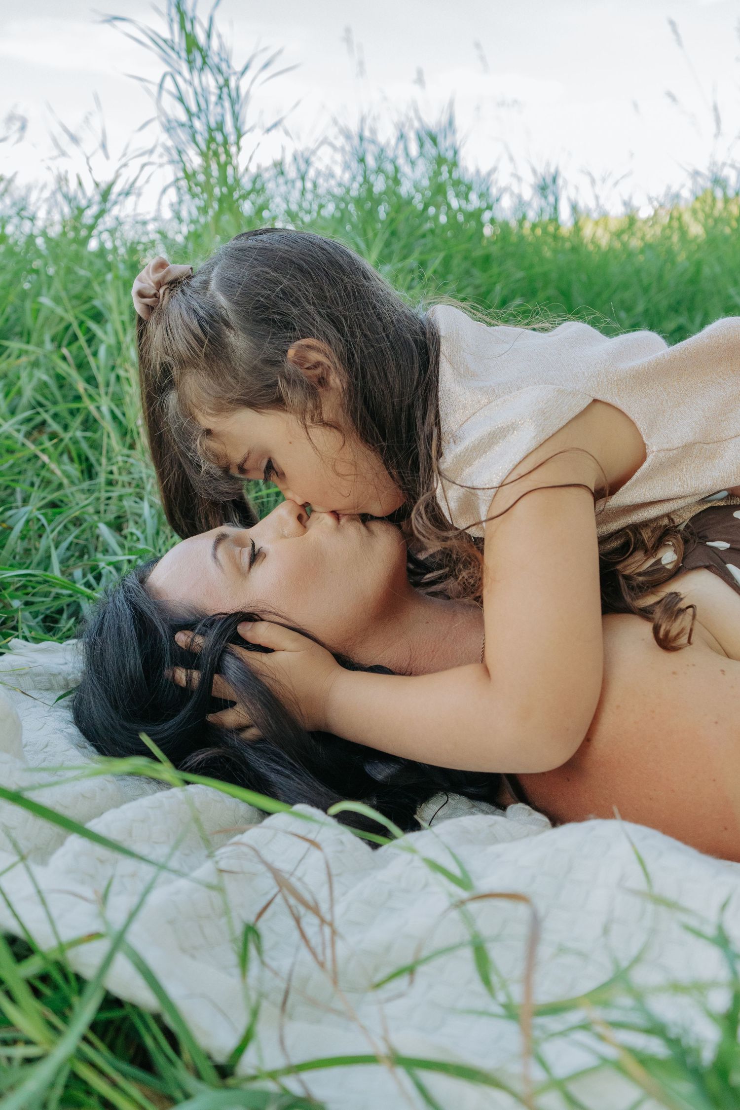 Little girl leaning in to kiss her mom's cheek as they lie on a blanket in the grass, sweet candid mother and daughter portrait