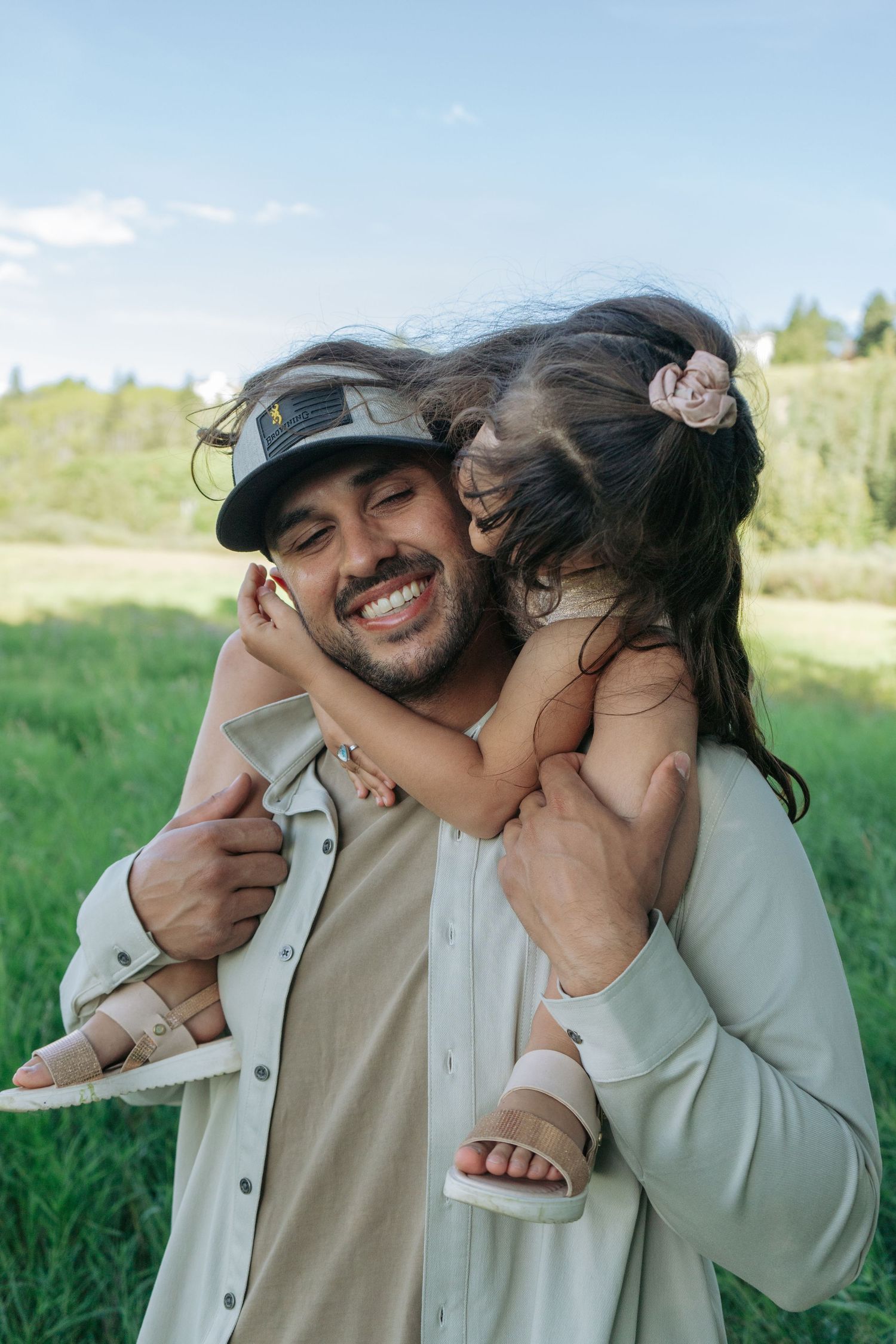 Dad in a ball cap smiling with his young daughter on his shoulders, laughing candid family portrait in a grassy field