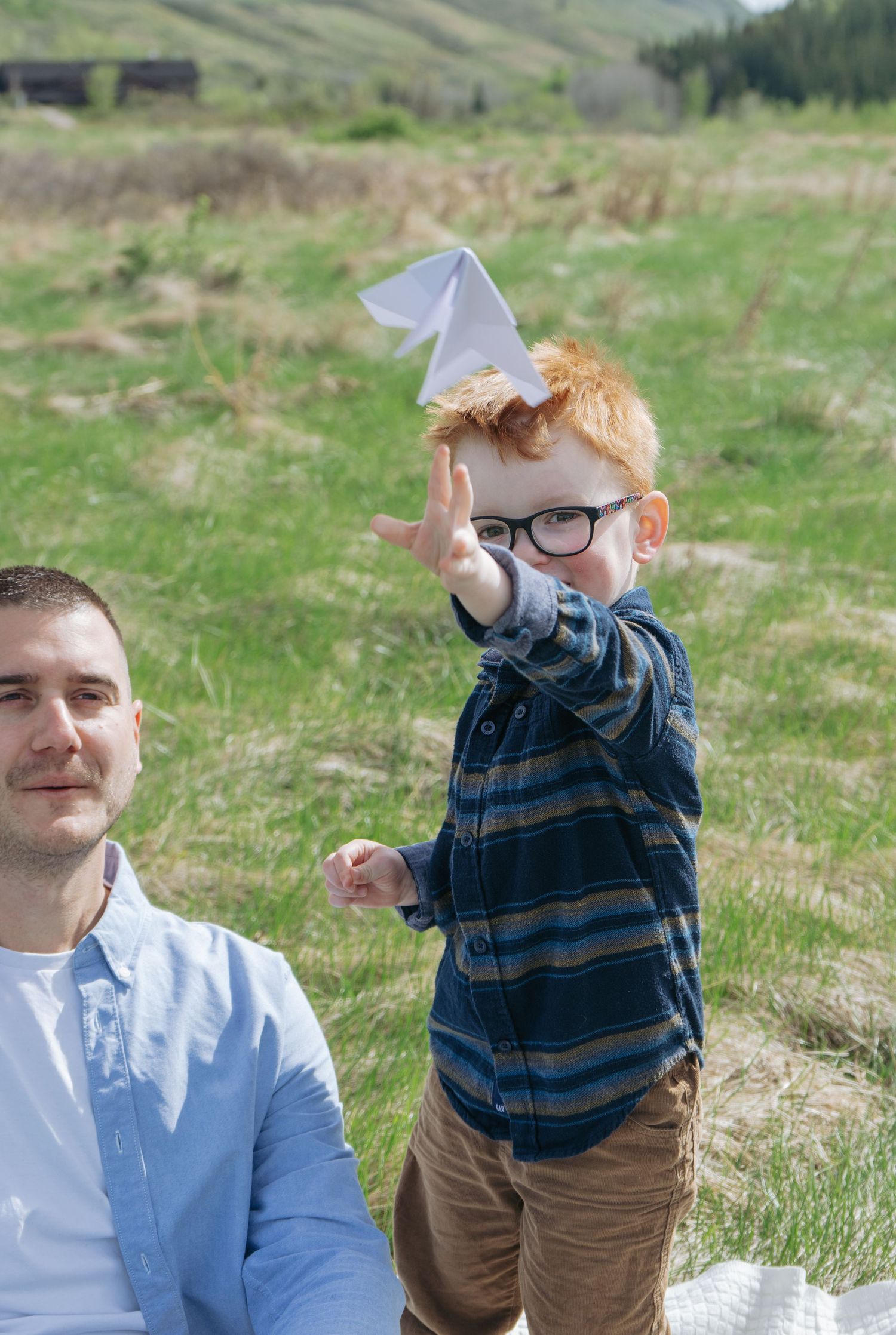 Dad smiling as red-haired toddler climbs on him outdoors, Cochrane family photography