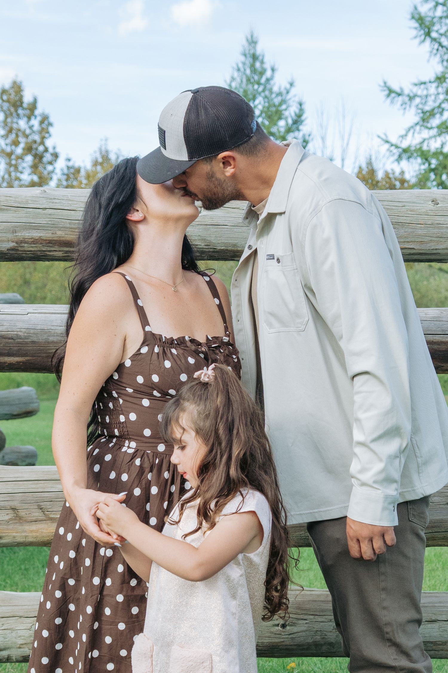 Mom and dad sharing a kiss in front of a rustic log fence while their daughter hugs mom's waist, romantic candid family portrait