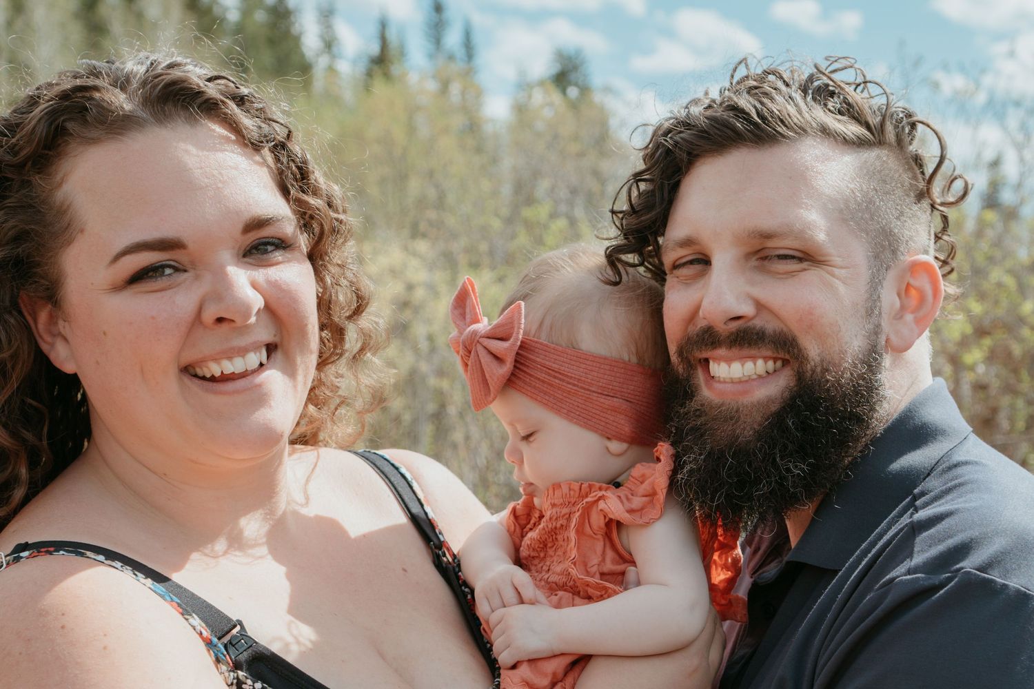 Smiling parents holding their baby girl outdoors with Alberta trees in the background, Cochrane family photography