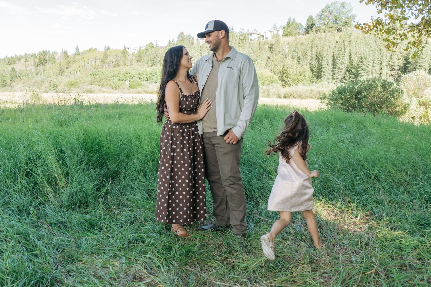 Mom and dad gazing at each other in a grassy field while their little girl runs past in a light dress, candid family photography