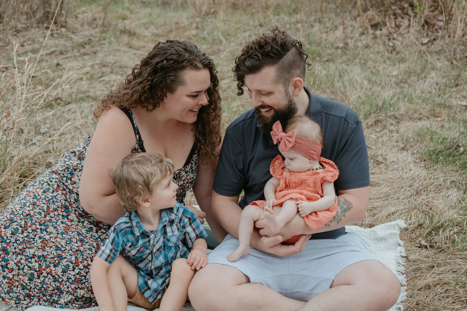 Family of four sitting together in a golden field admiring their newborn, Alberta family photography