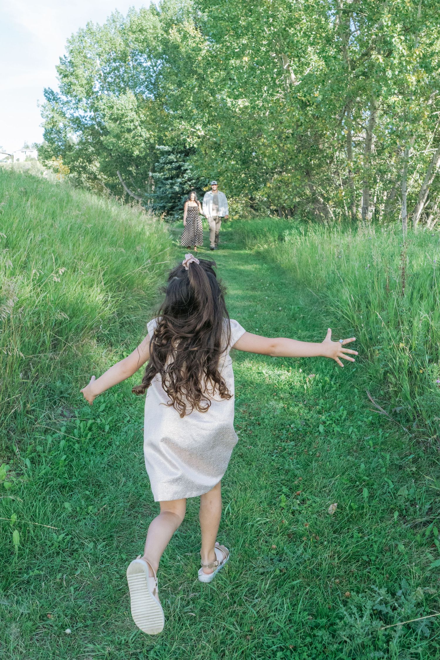 Young girl running down a mown path through tall green grass with arms outstretched, parents walking in the distance