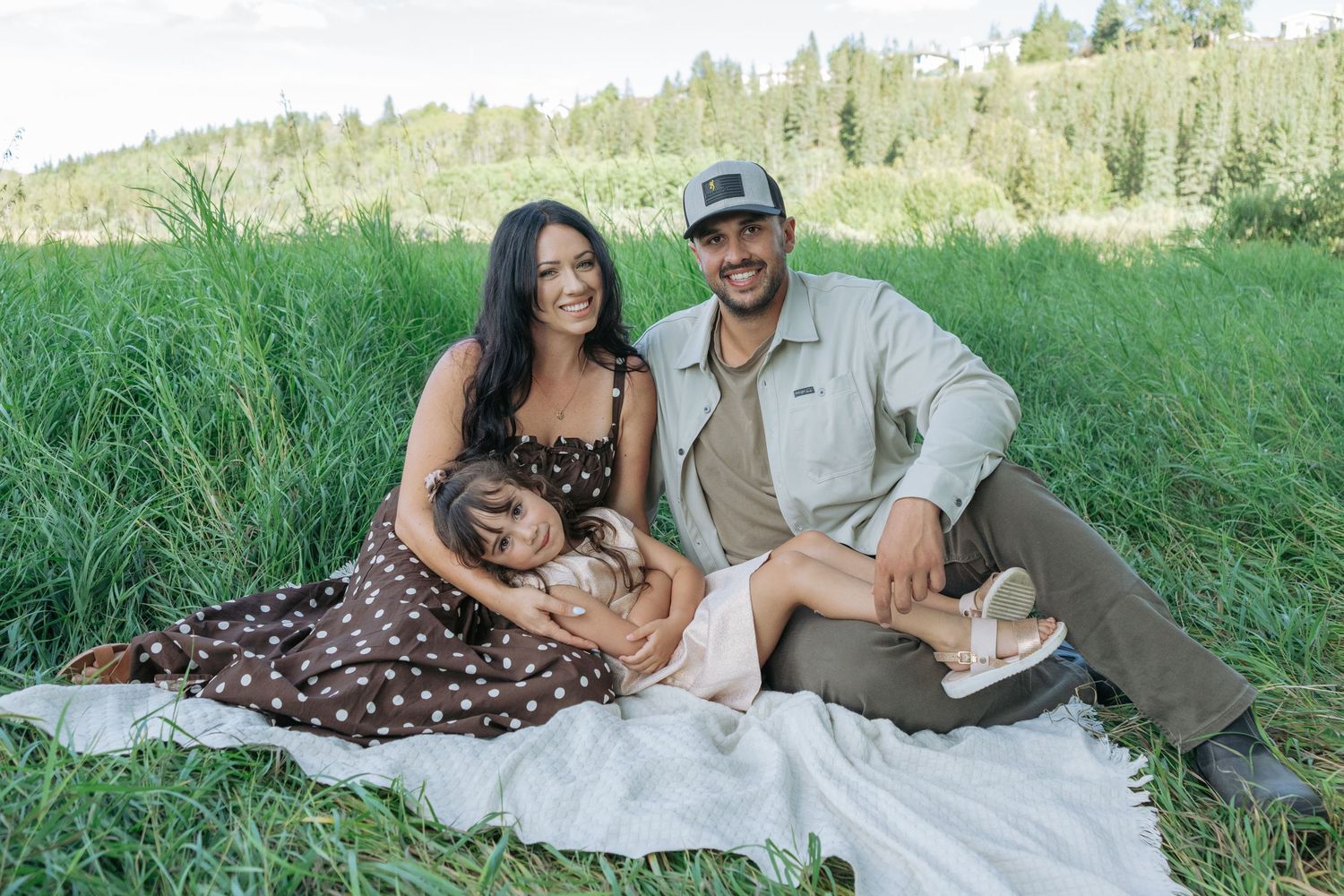 Mom, dad, and daughter relaxing on a blanket in tall green grass, daughter curled up in mom's lap, family photography in the Bow Valley