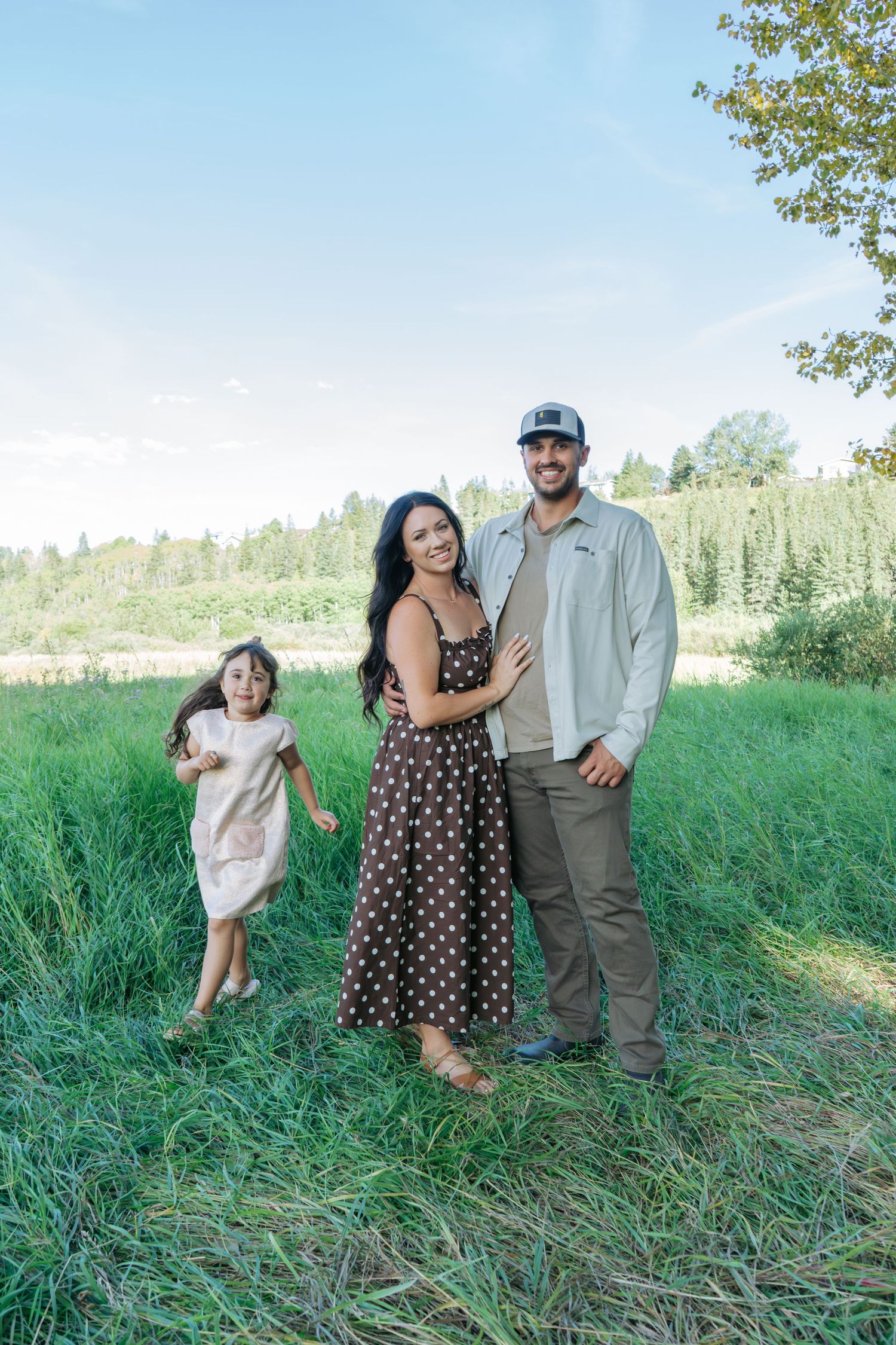 Mom and dad standing close together in a sunny meadow while their little girl stands off to the side, bright outdoor family photography
