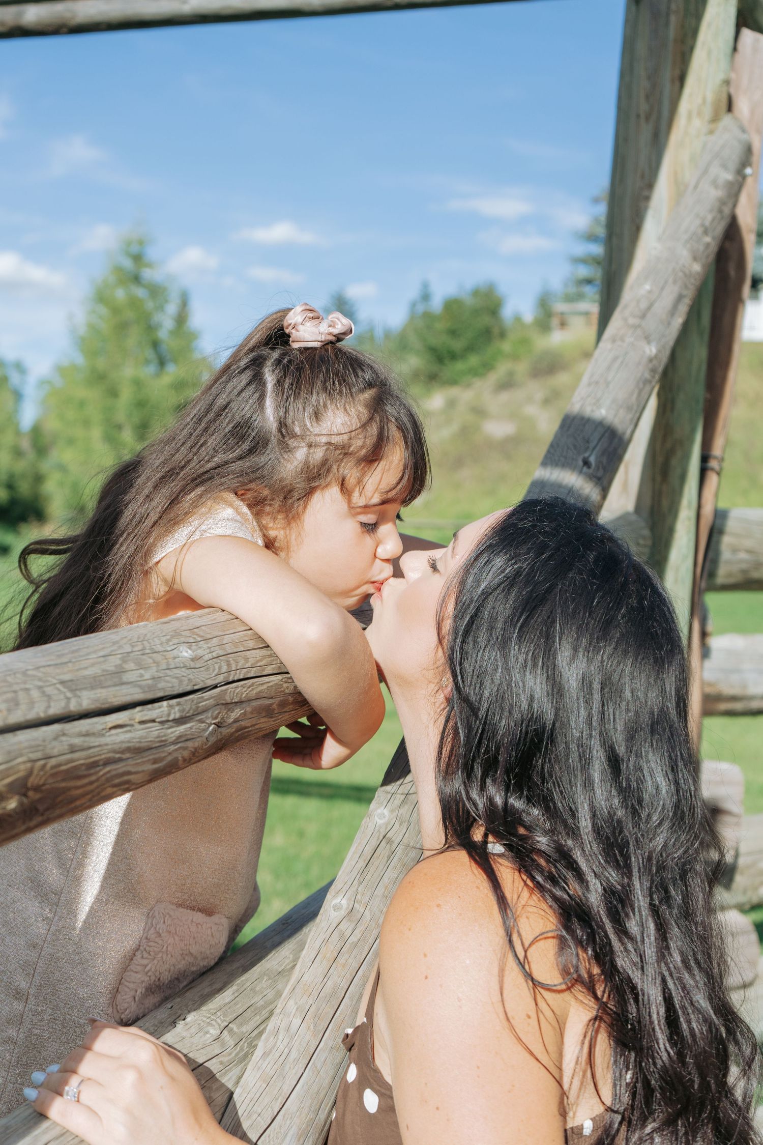 Little girl leaning over a rustic wooden fence to kiss her mom, sweet candid mother and daughter moment outdoors