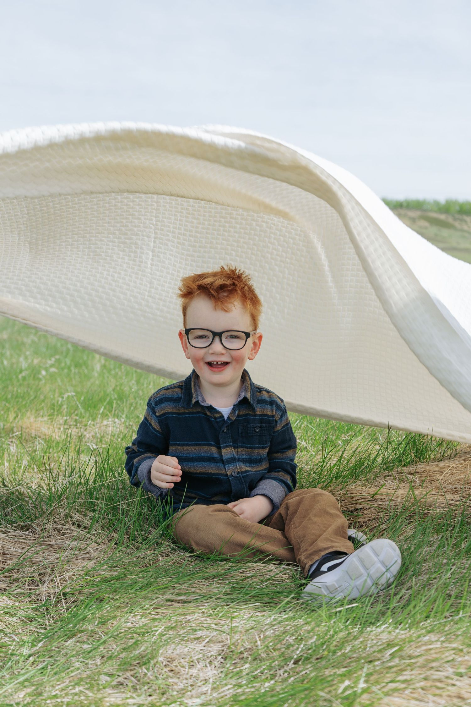 Red-haired toddler sitting in the grass under a blanket tent, candid childhood moment