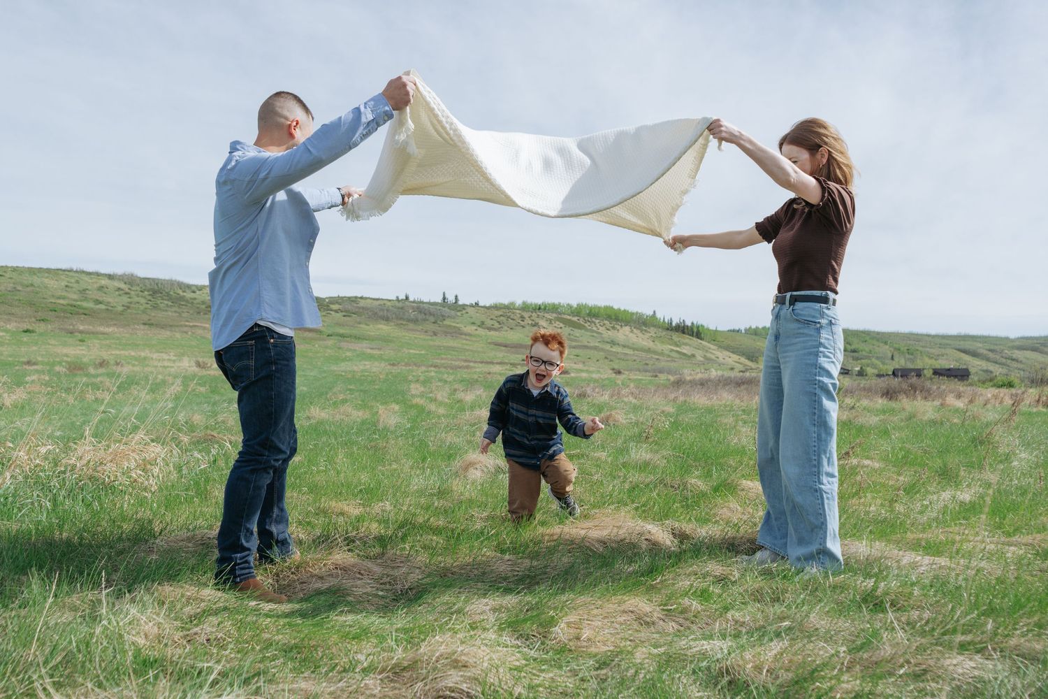 Parents lifting a blanket over a toddler in an open field, playful Cochrane family portrait