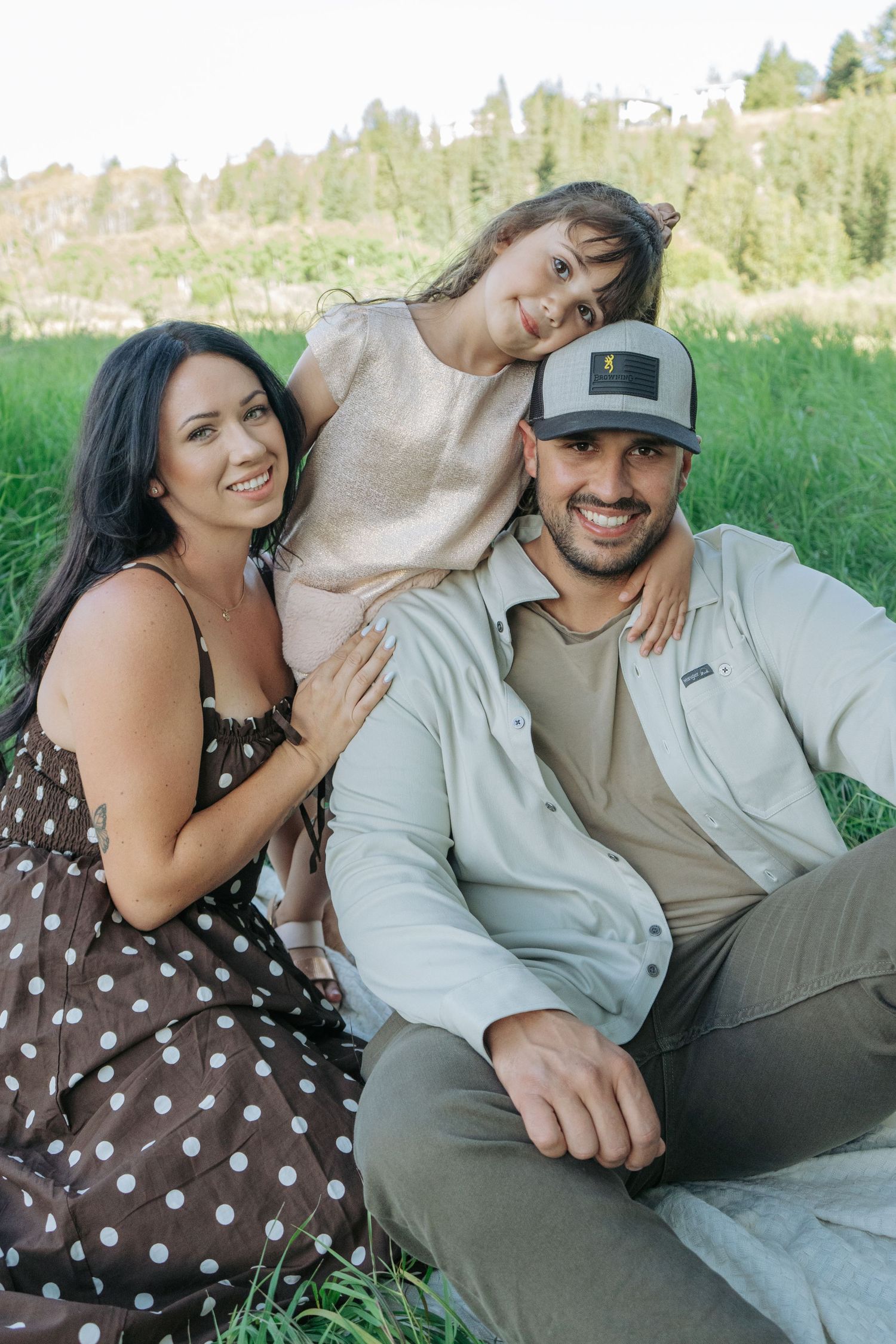 Mom, dad, and daughter sitting close together in the grass, all smiling at the camera, warm natural family portrait near Cochrane