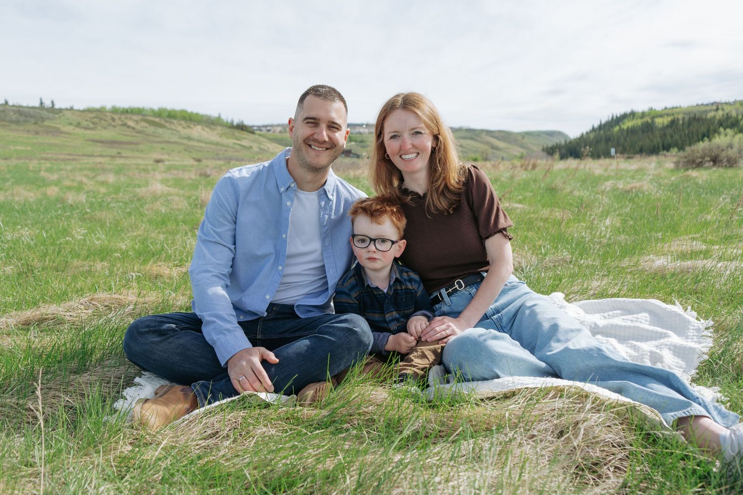 Family of three sitting together on a grassy hillside, Cochrane family portrait by Maddock Studios