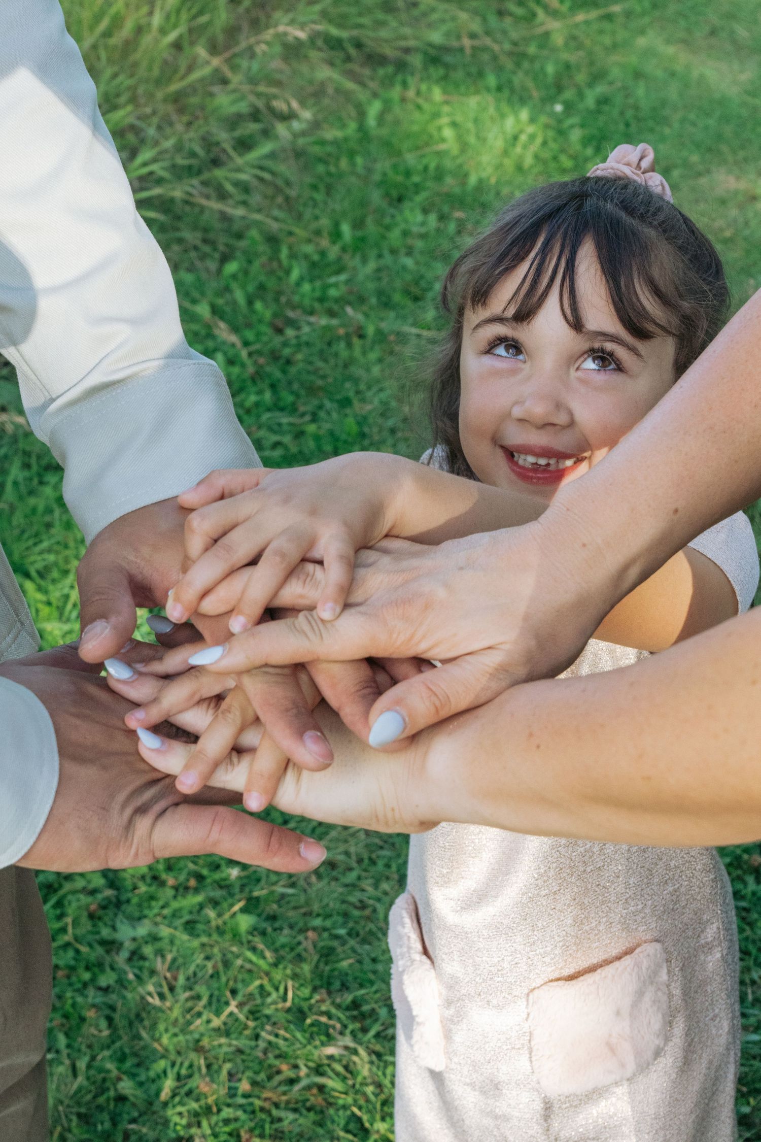 Close-up of a little girl smiling up as the family stacks their hands together in the grass, playful family portrait detail shot