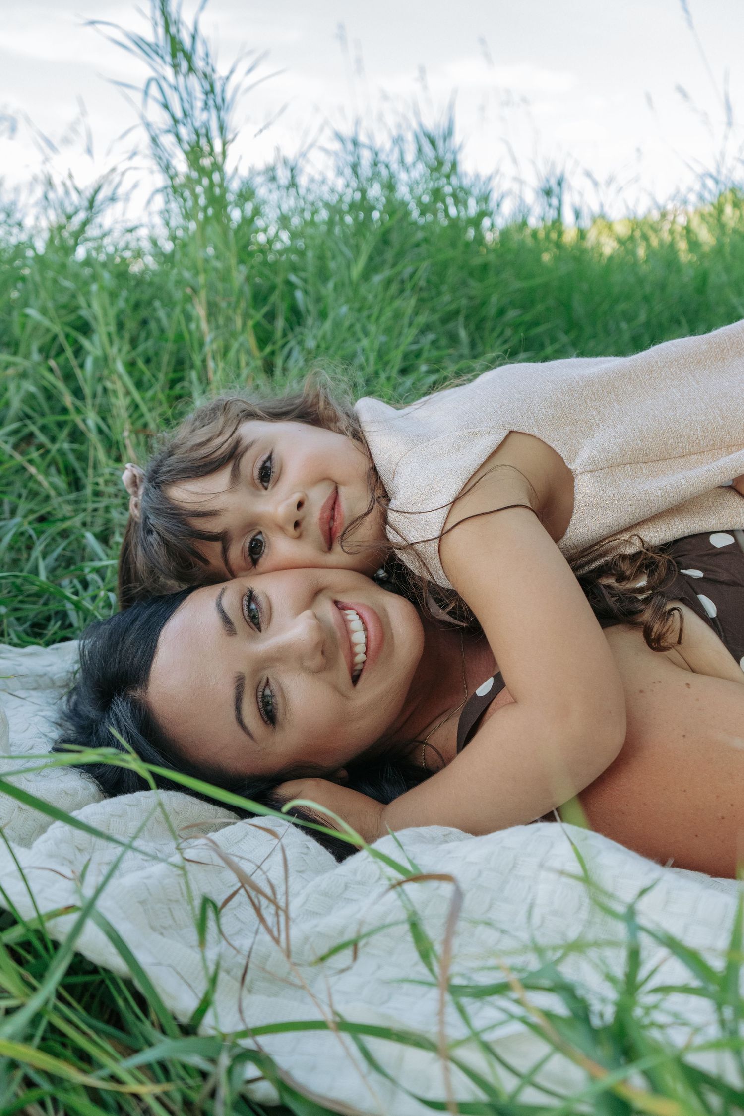 Mom and daughter lying on a blanket in the grass, smiling at the camera, natural outdoor family portrait