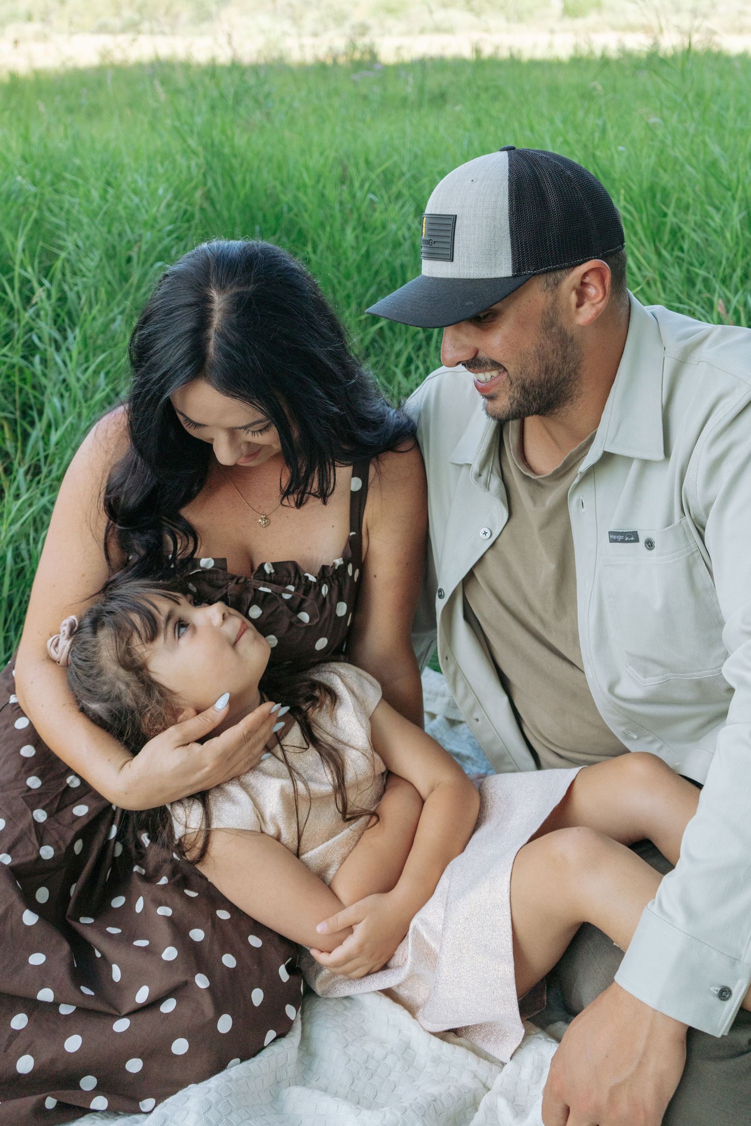 Mom and dad sitting close in the grass, cuddling their little girl, soft candid family portrait