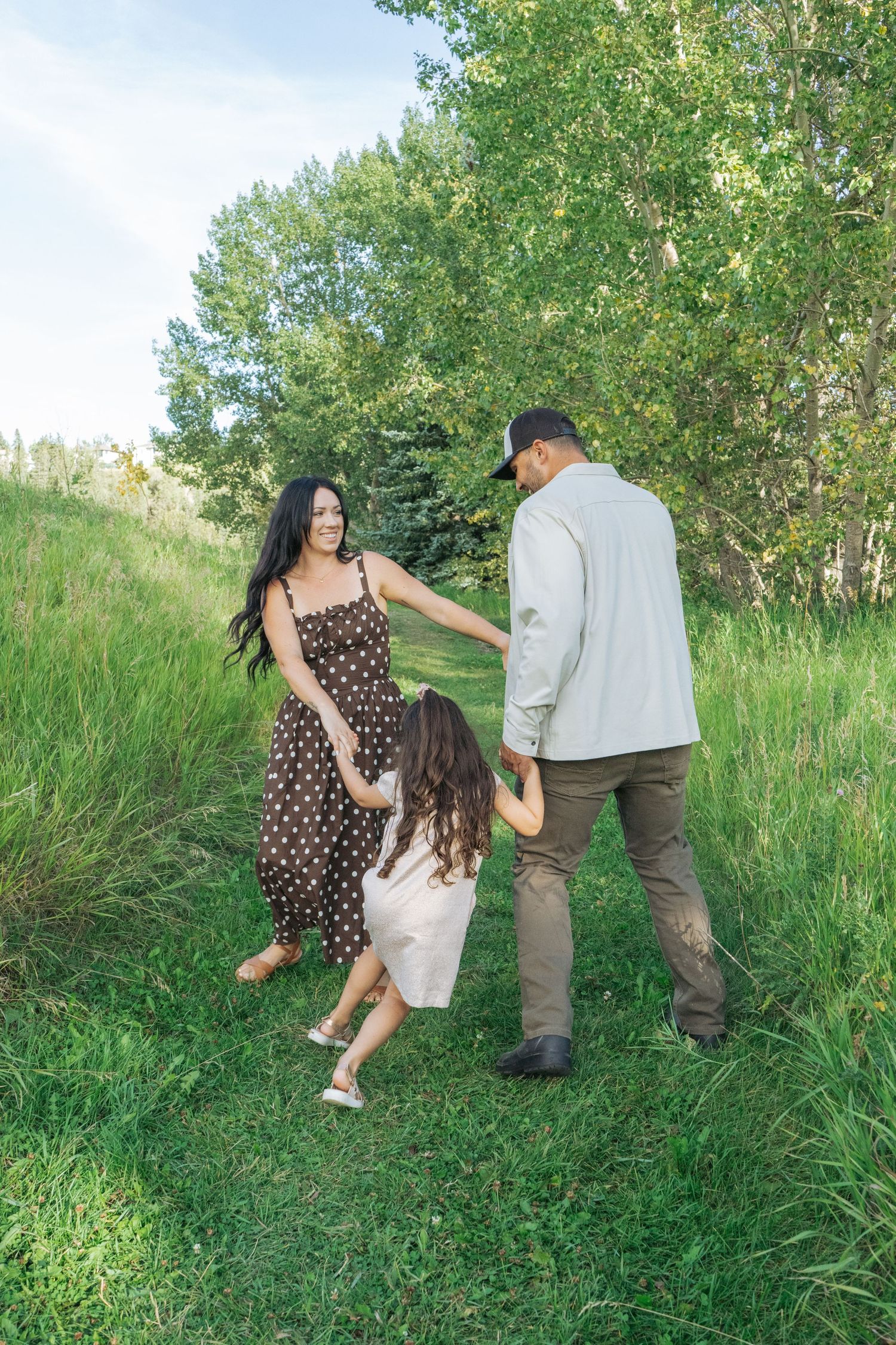 Parents and daughter walking hand in hand down a grassy path lined with trees, relaxed outdoor family session in the Bow Valley
