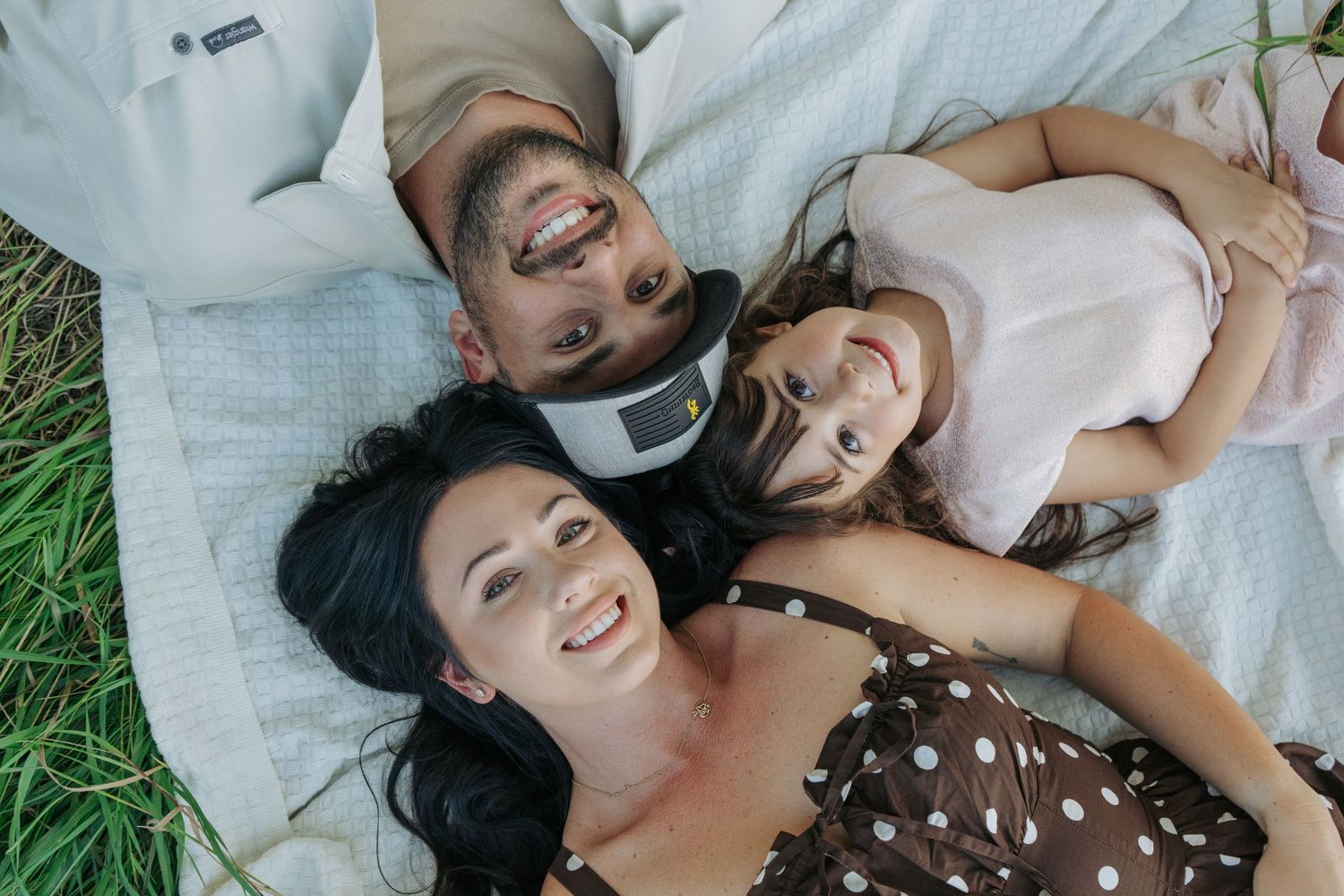 Overhead shot of mom, dad, and daughter lying on a blanket in the grass with their heads together, all smiling up at the camera