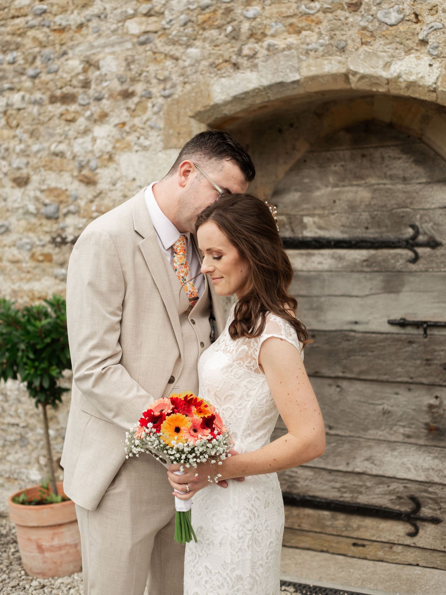 A Bride & Groom embrace against a rustic stone wall with a vibrant orange and pink floral bouquet.