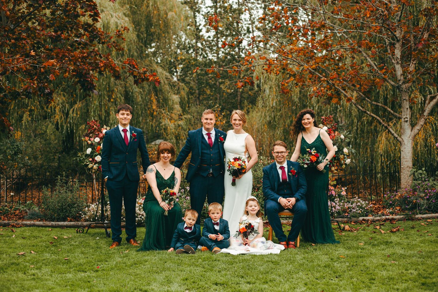 Wedding party in navy blue attire poses on the grass lawn of Rumbold Farm, with autumn foliage backdrop during an Autumn ceremony.