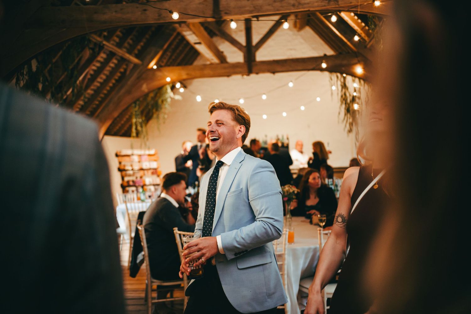 A wedding guest in a light blue suit jacket mingles at the wedding breakfast with string lights and wooden beams overhead @ Southstoke Barn.