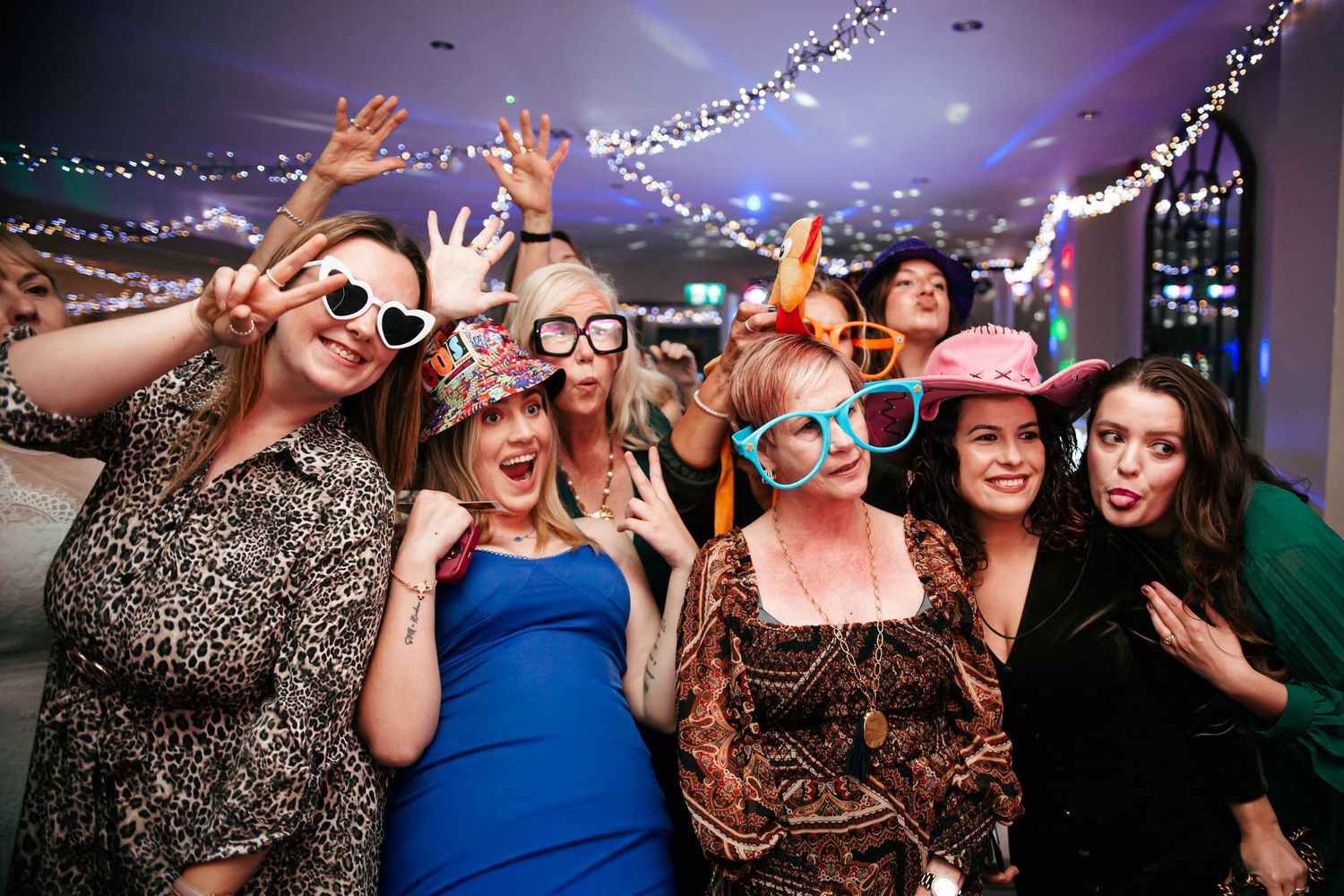 Group of party guests wearing colorful sunglasses and fun props while posing and dancing during the evening.