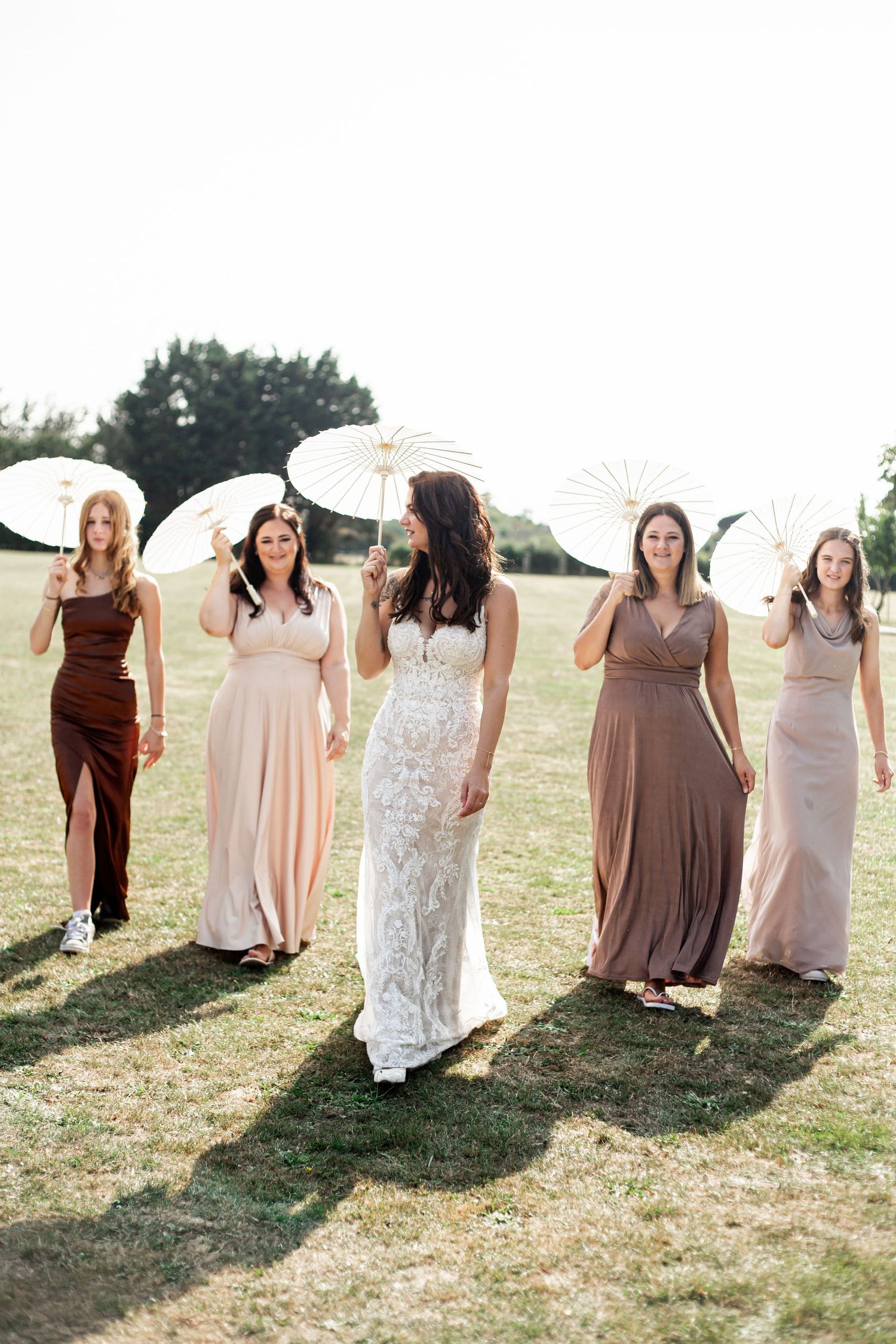 The bridal party holding white parasols pose together on the grassy field during a sunny day. Barnham Community Field, West Sussex.