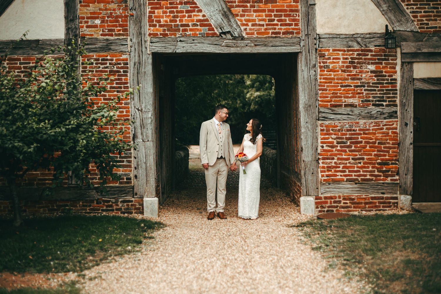 A wedding couple stands in the doorway of a rustic brick barn building with a gravel path.