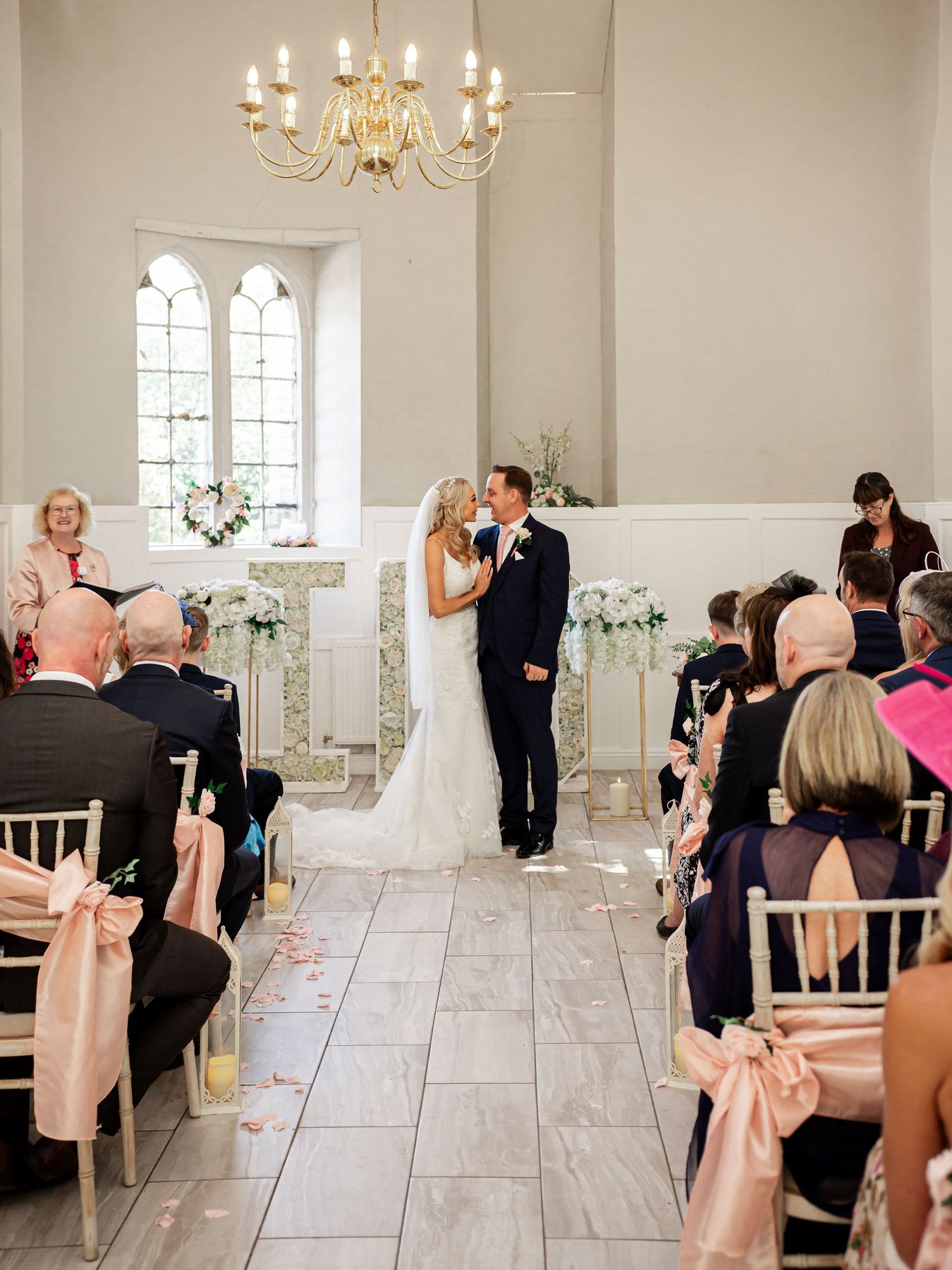 A wedding ceremony takes place in the elegant white chapel of Goring Castle with arched windows and a crystal chandelier.