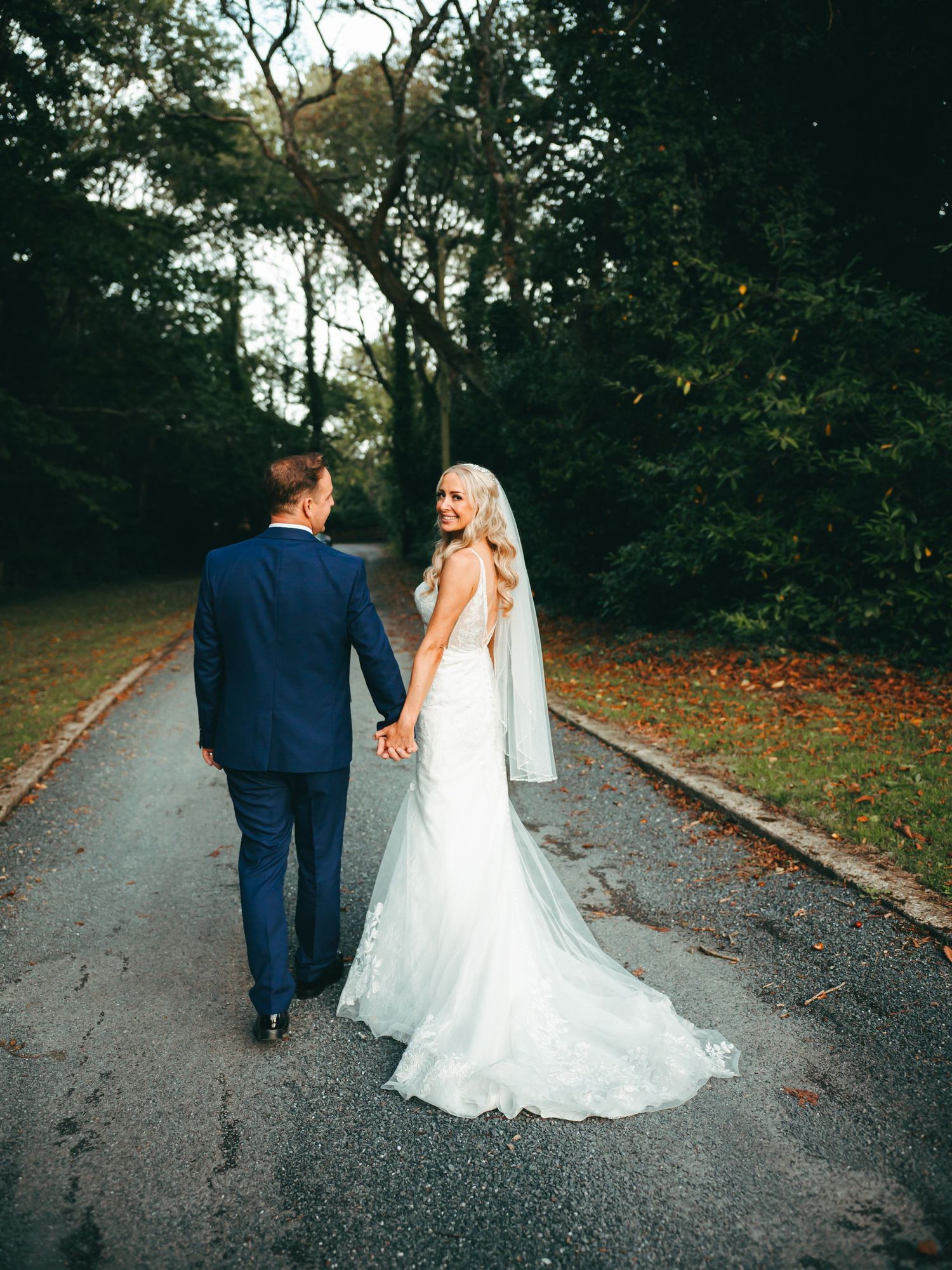 A Bride & Groom walk down a tree-lined path with scattered autumn leaves during an Autumn wedding.